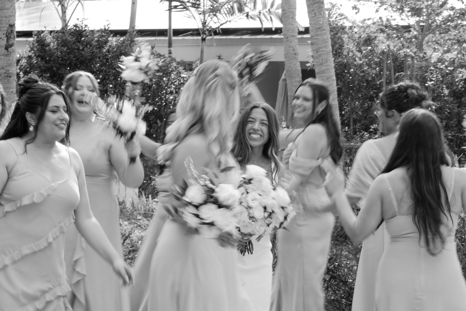 Black and white photo of bride with bridesmaids in light dresses laughing and celebrating outdoors.