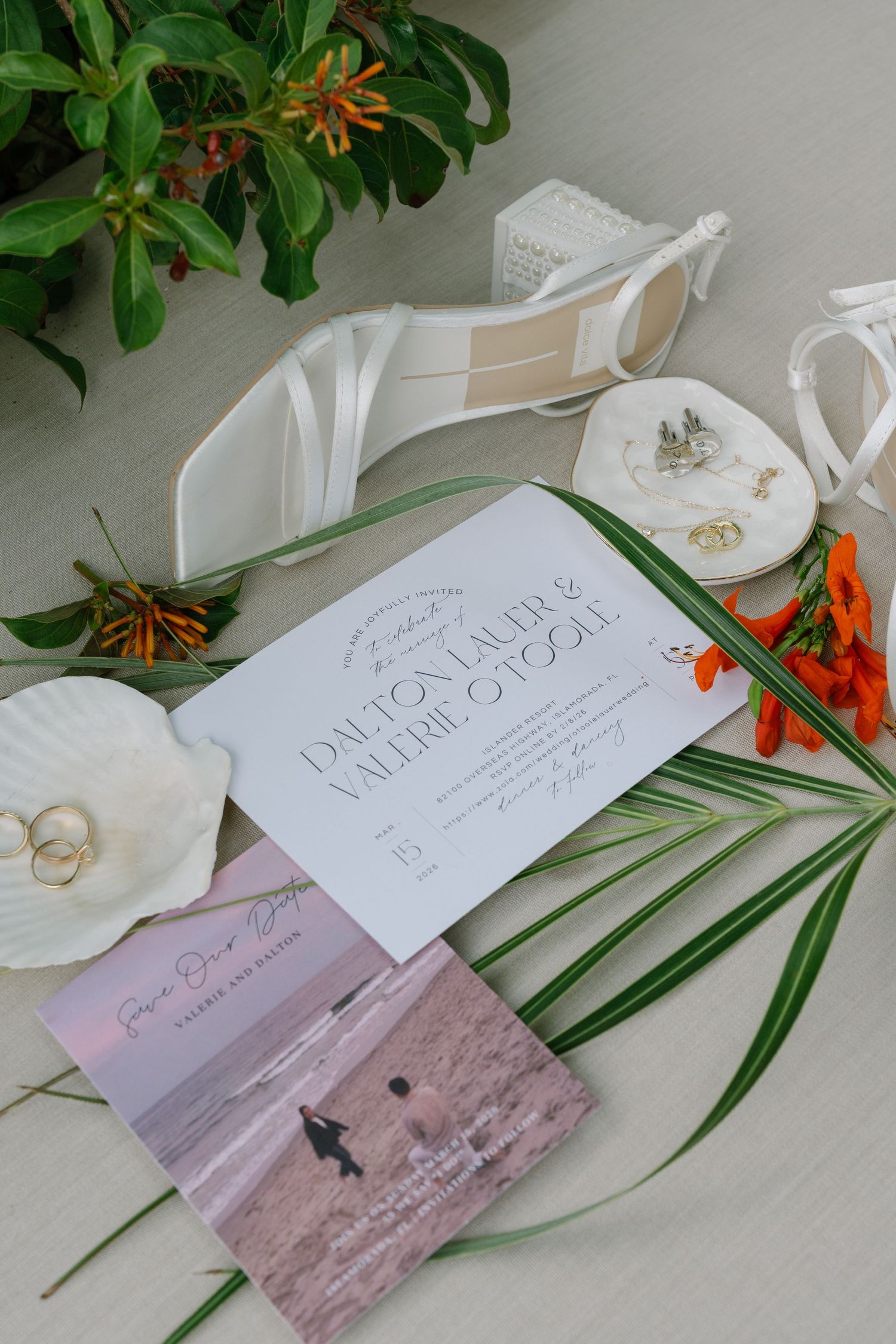 Wedding invitation suite with white heels, rings, and tropical flowers on a neutral surface.