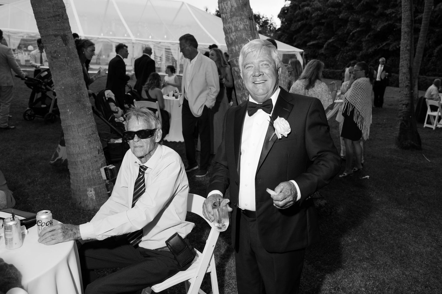 Two men in tuxedos smiling at an outdoor wedding reception in black and white photo.
