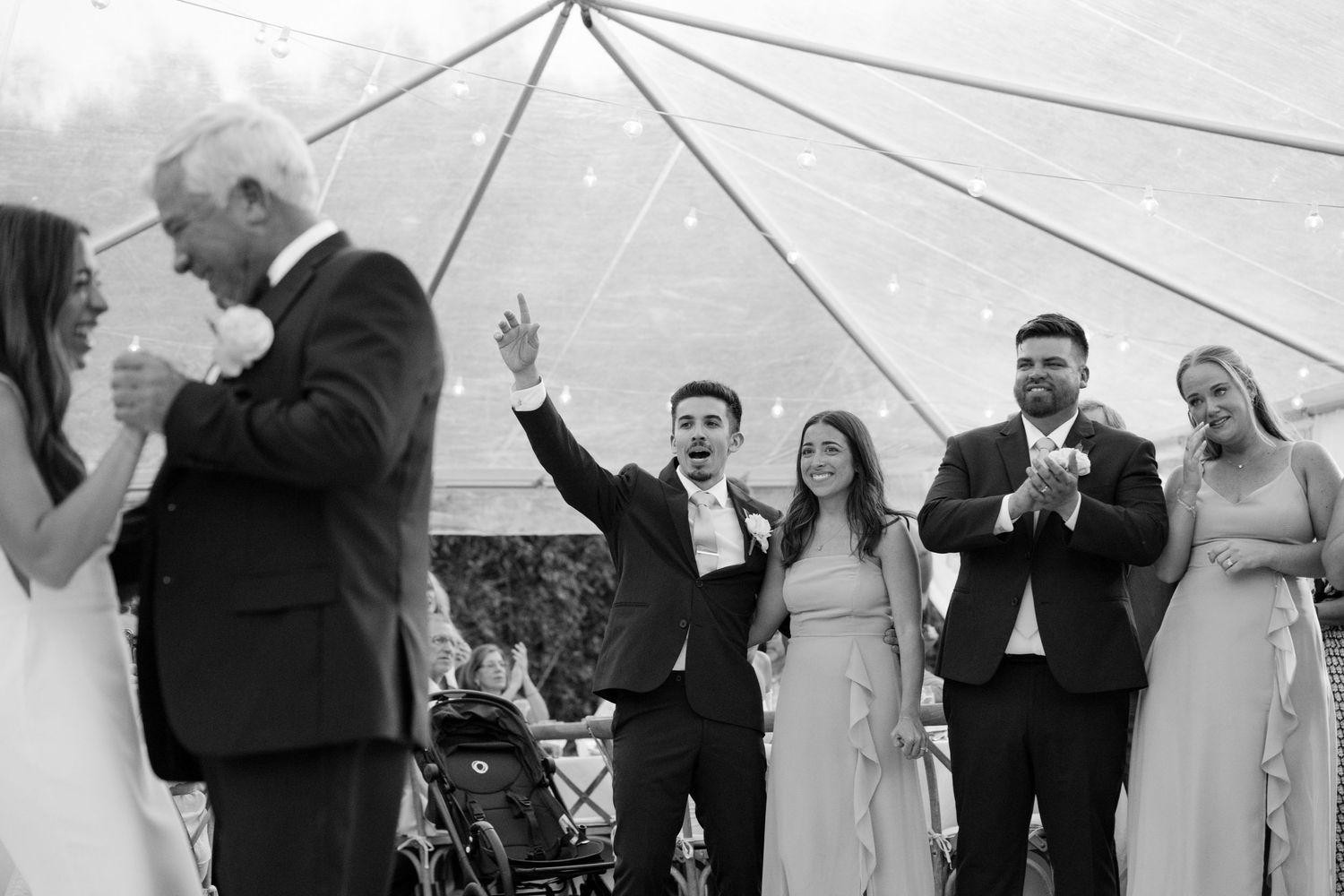 Black and white photo of groom celebrating with raised arm surrounded by wedding guests under a tent.