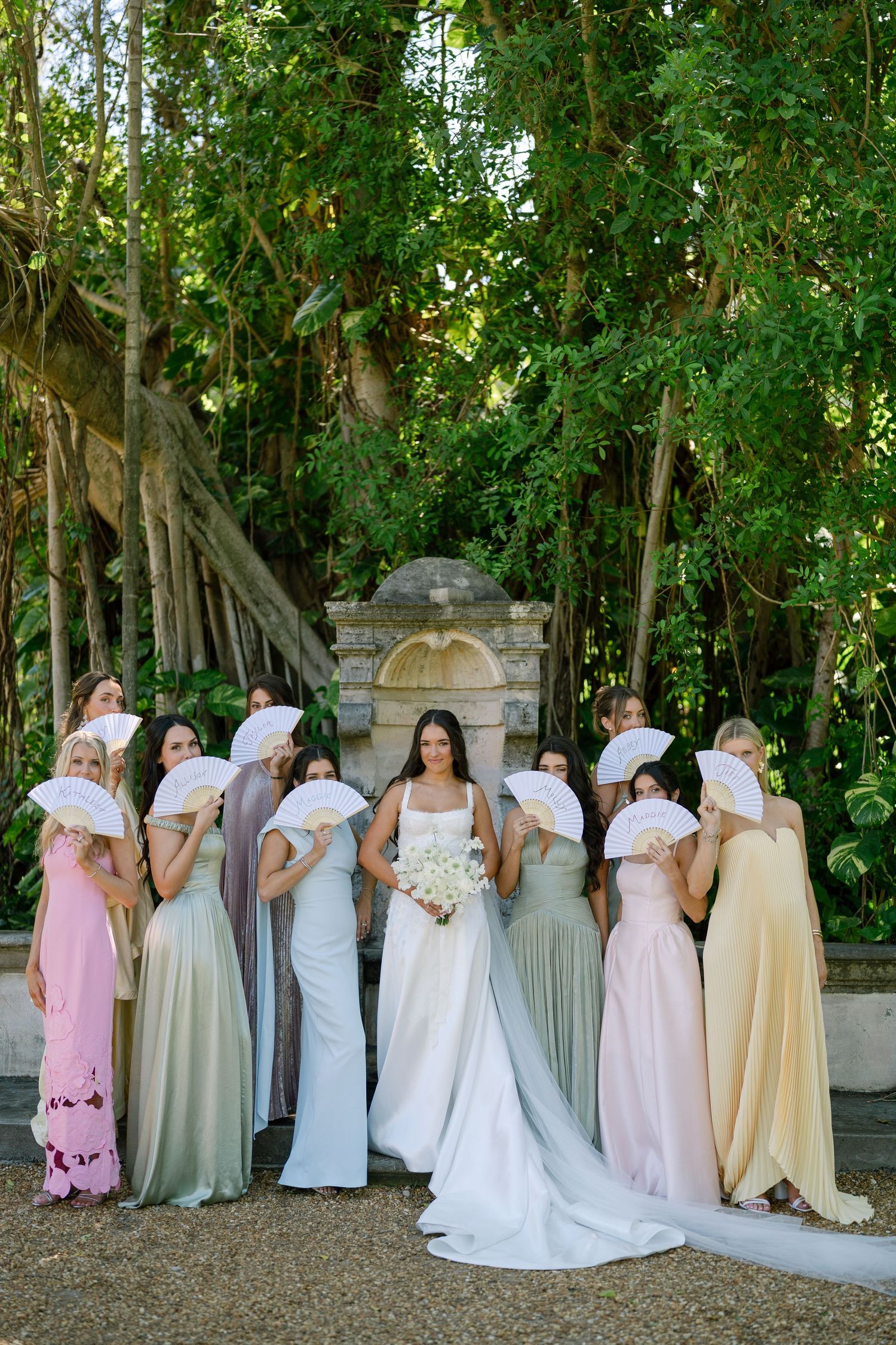 Bride in white gown poses with bridesmaids in pastel dresses holding white fans in lush garden setting.