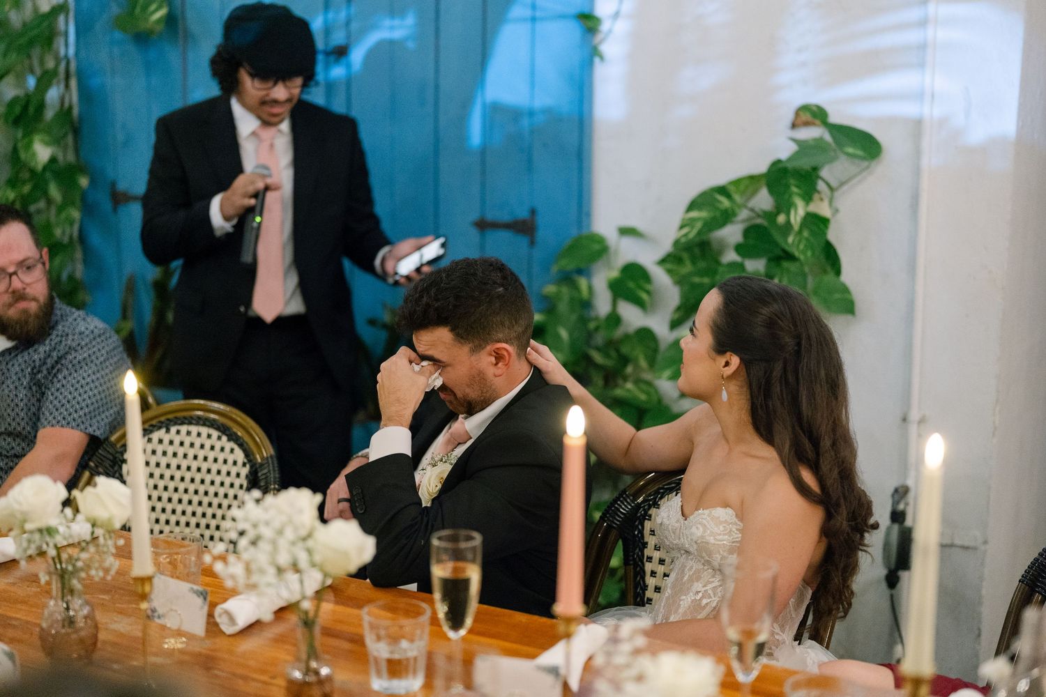 Bride touches grooms head at candlelit reception table while man speaks into microphone behind them.