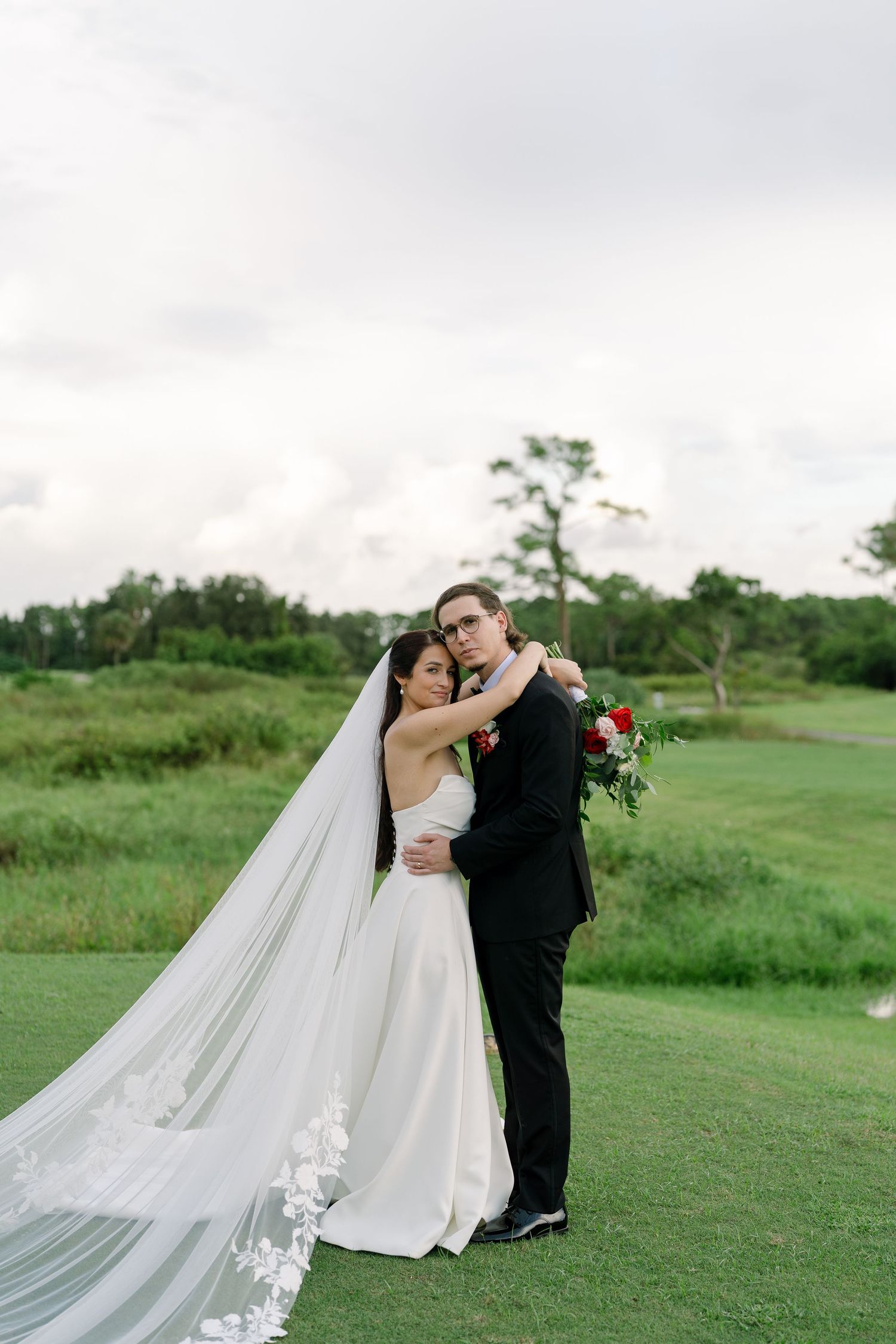 Bride and groom embrace on lush green golf course, her long lace veil trailing, holding red bouquet under cloudy sky.