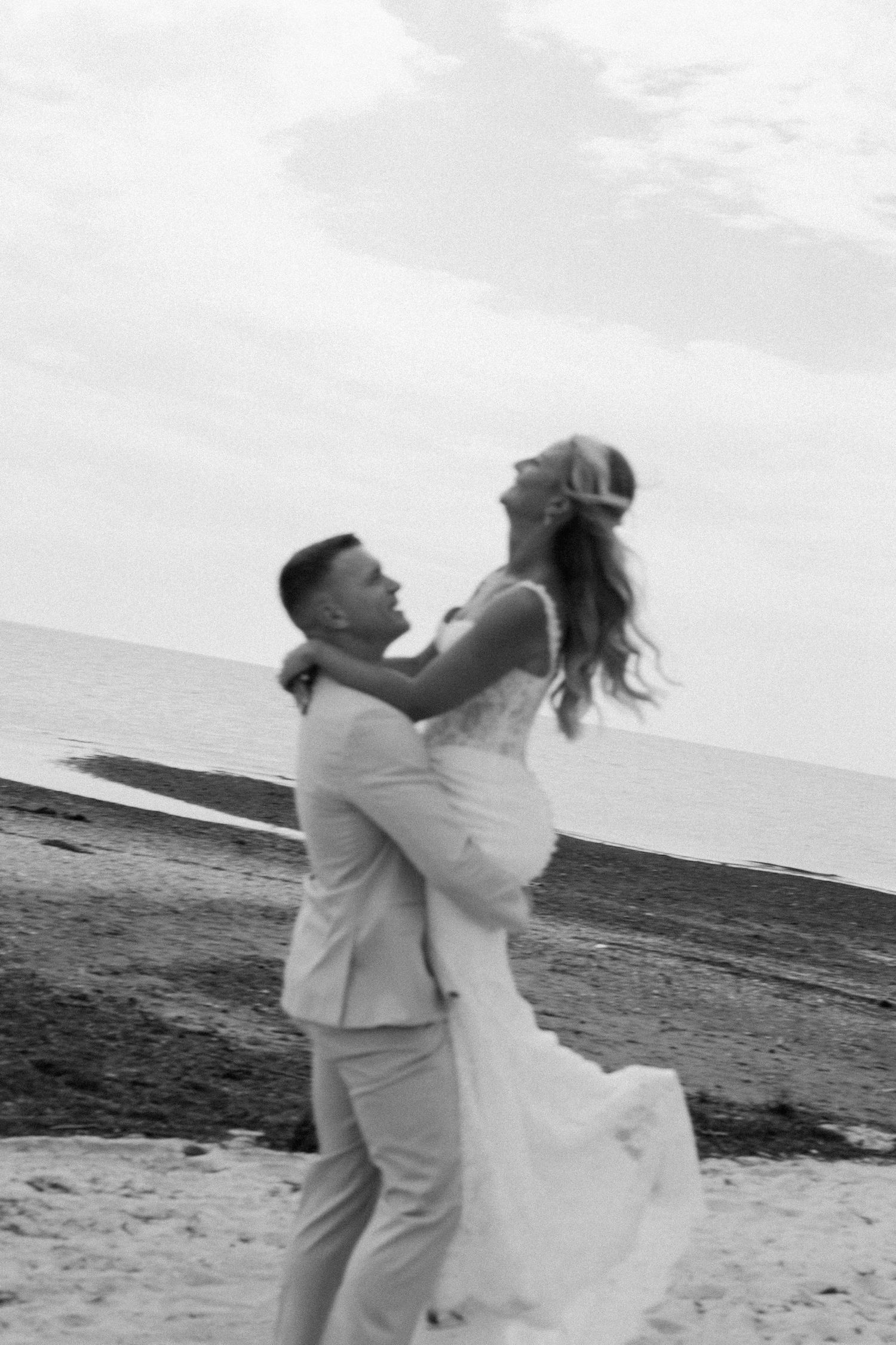 Black and white beach wedding photo of groom lifting bride in white gown, ocean backdrop, joyful romantic moment.