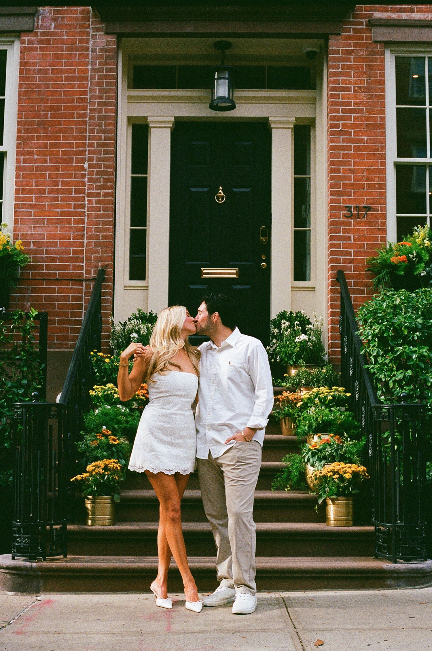 Couple kissing in white outfits on brick townhouse steps with yellow mums and lush greenery.