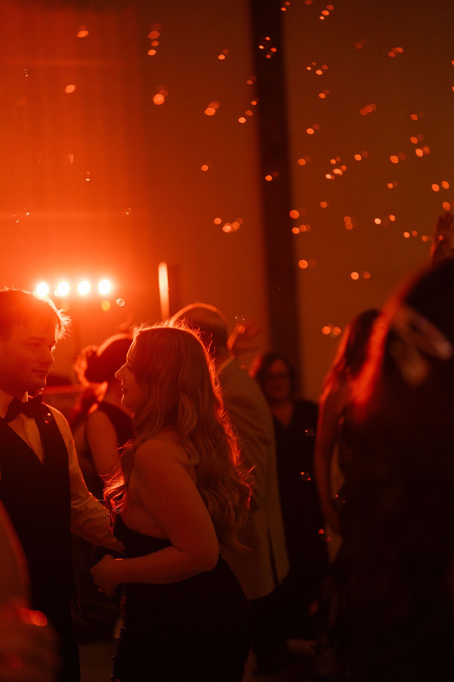 A crowd of people gather in a dimly lit room, with bright lights and colorful decorations visible in the background.