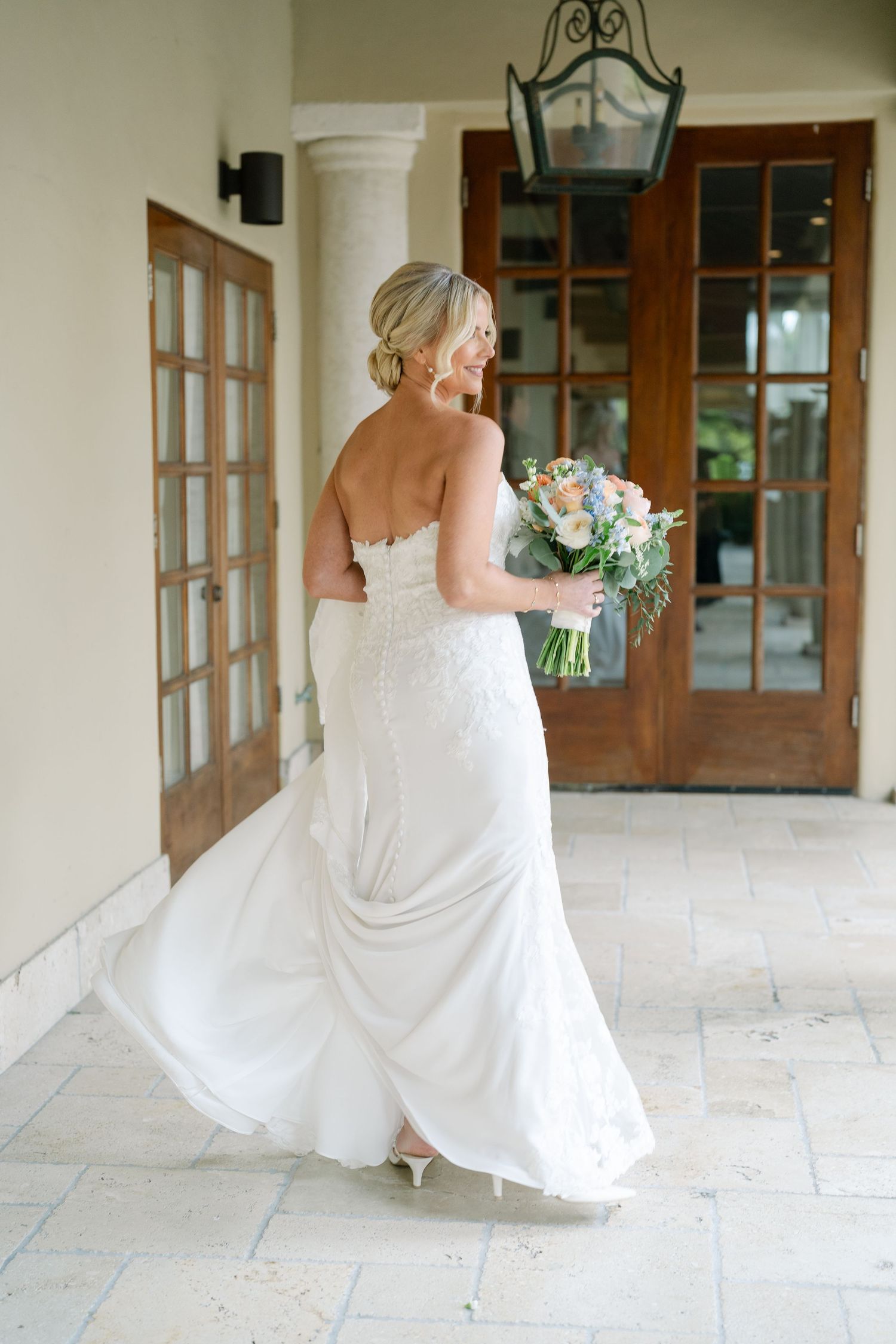 A bride in a flowing white dress holds a bouquet while standing on a porch with French doors.