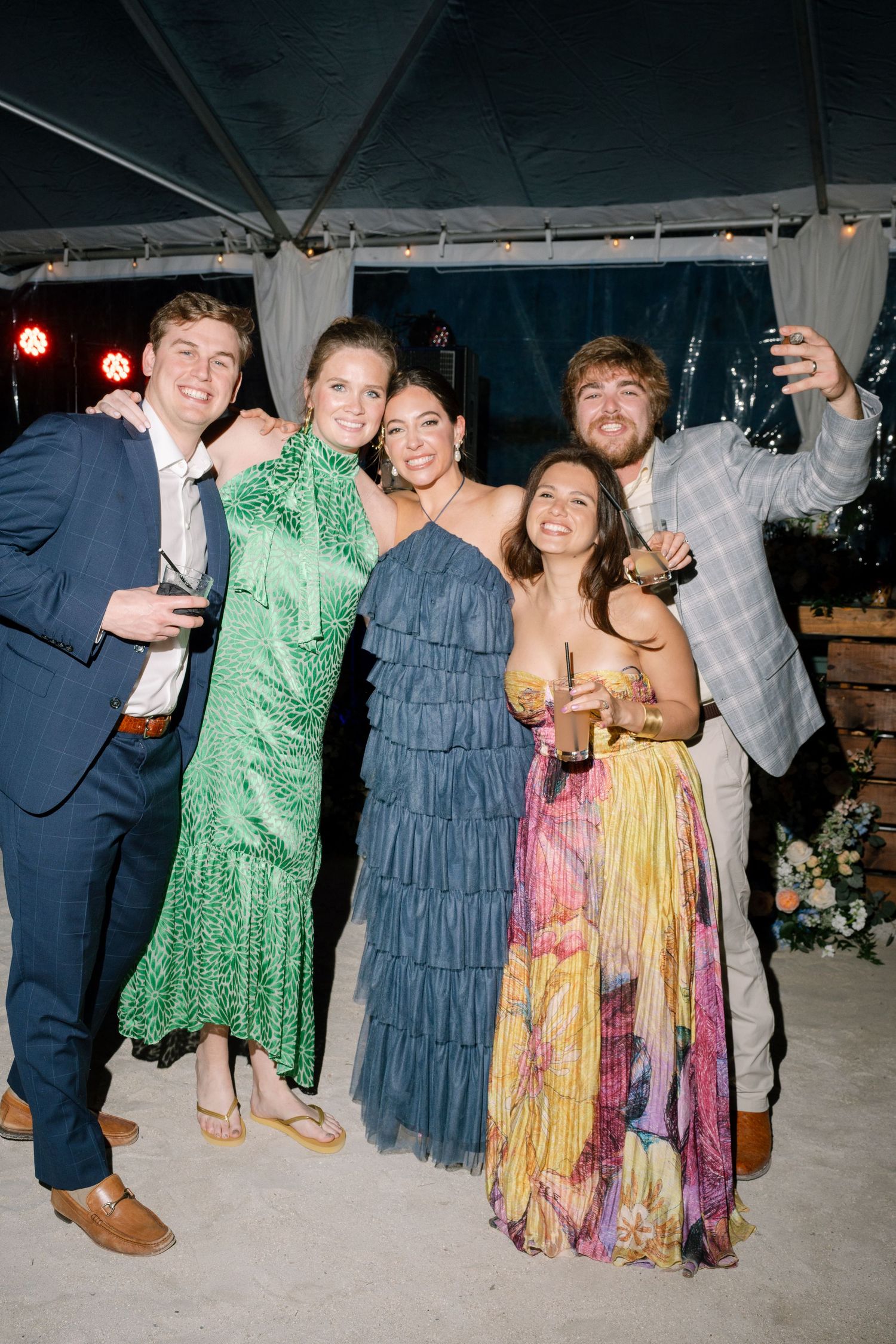 Party guests in colorful dresses dancing and posing together under a tent at night.