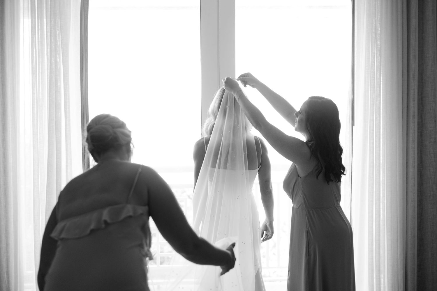 A silhouetted scene of bridal preparations by a window as a veil is being adjusted in soft natural light.