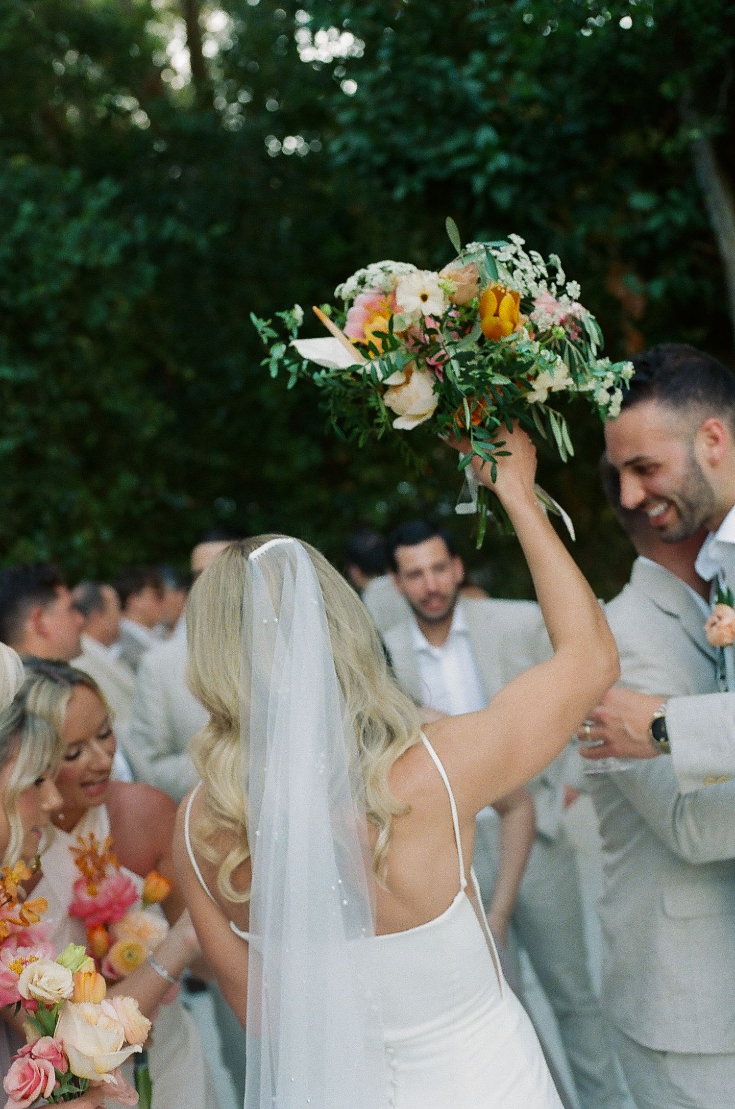 Wedding party members in light colored attire celebrate during an outdoor garden ceremony with floral bouquets.