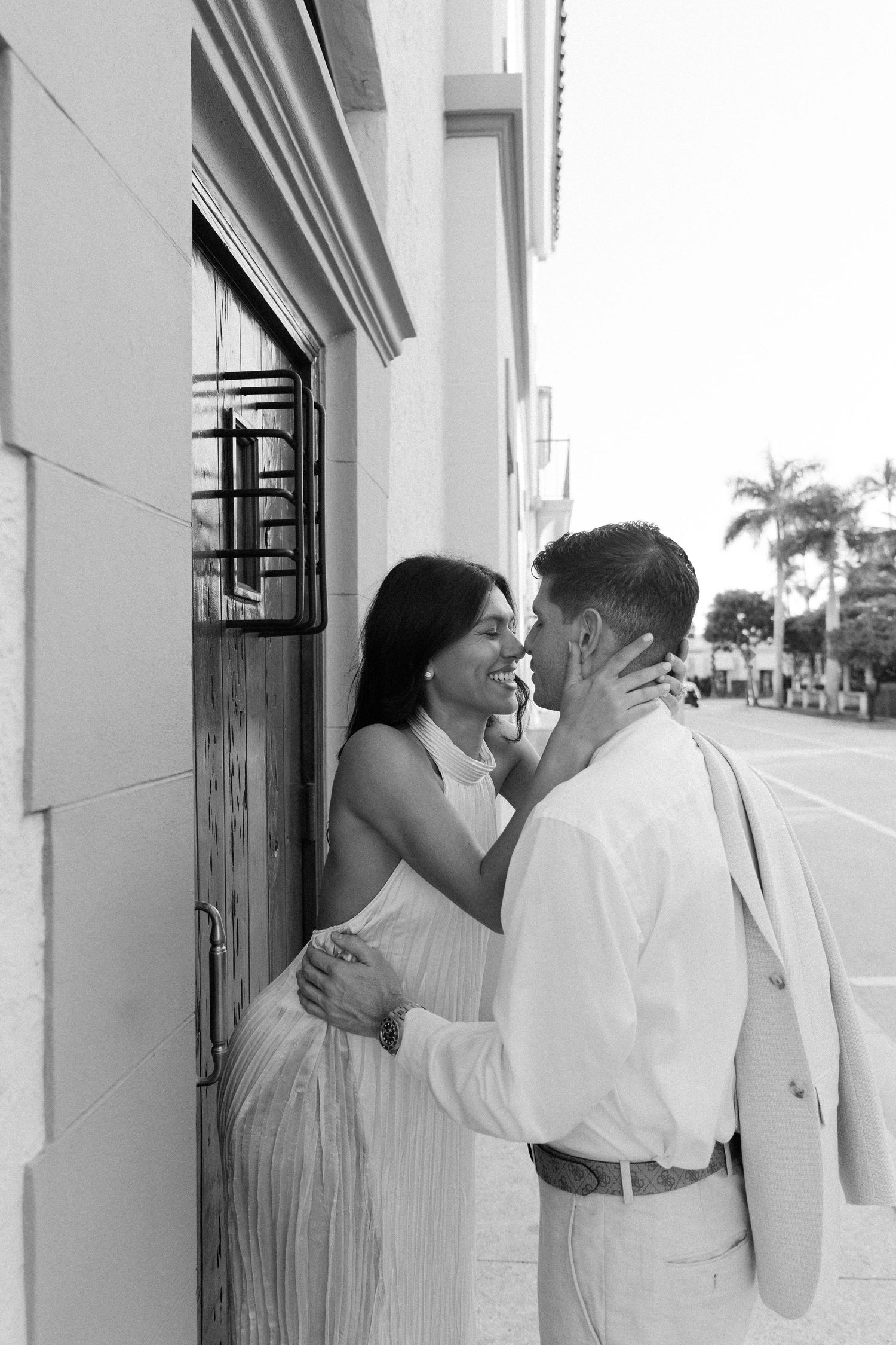 A romantic black and white photo of a couple embracing and smiling together on an urban street with palm trees.