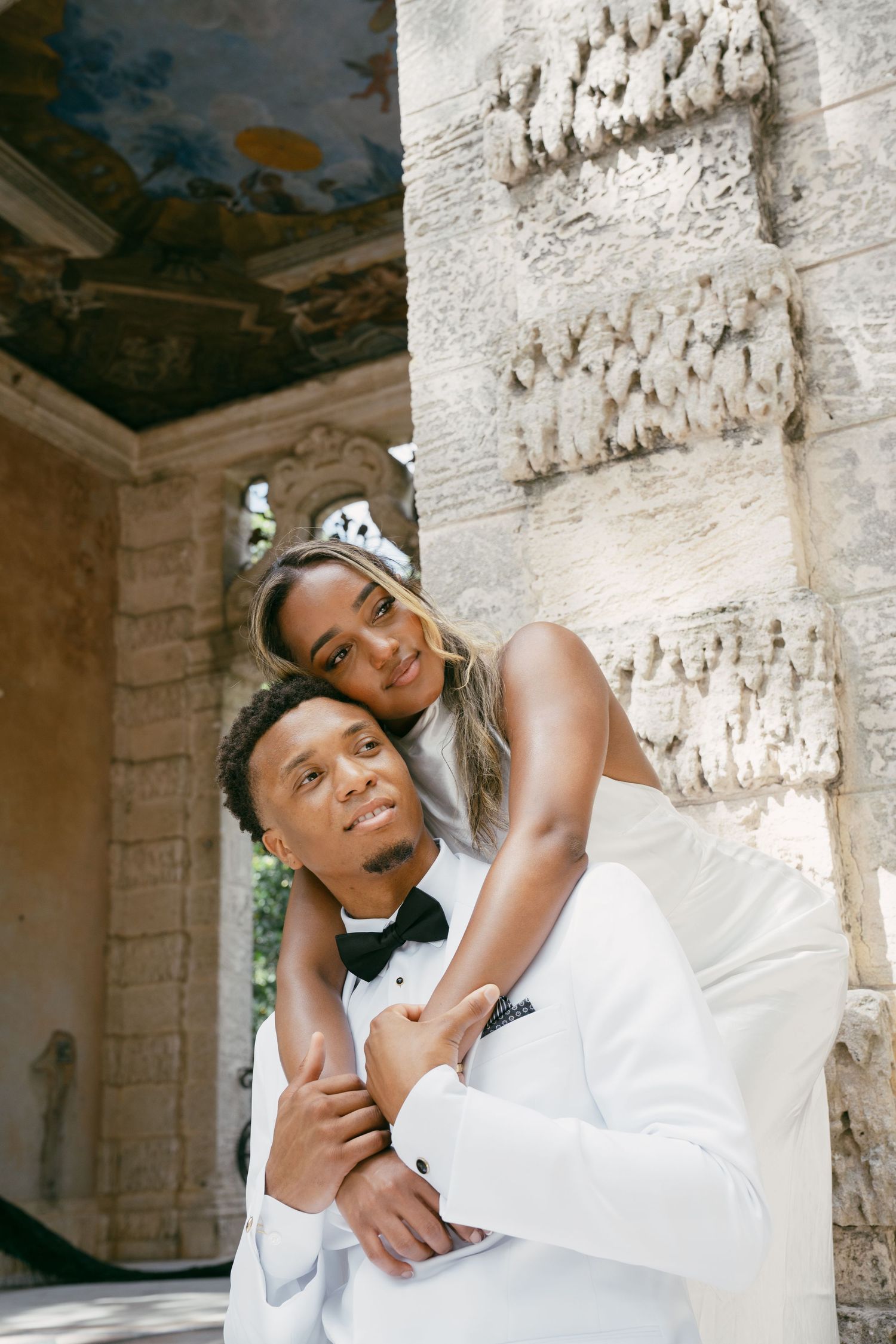 A wedding couple embraces while posing in front of a stone wall with decorative architectural details.