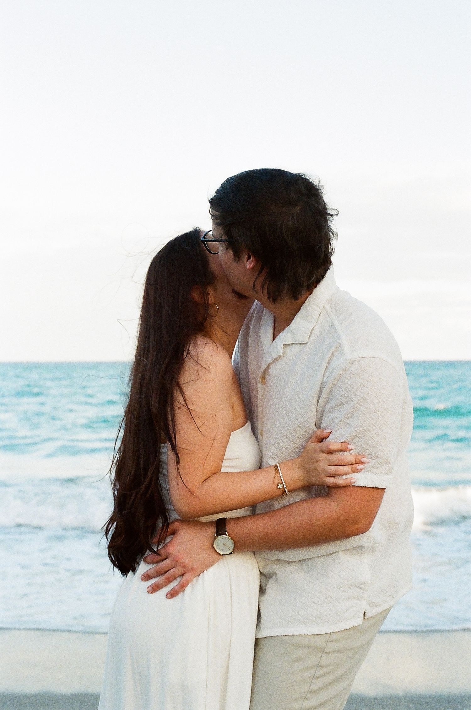 A tender moment between a couple in white outfits embracing on a beach with blue waters in the background.