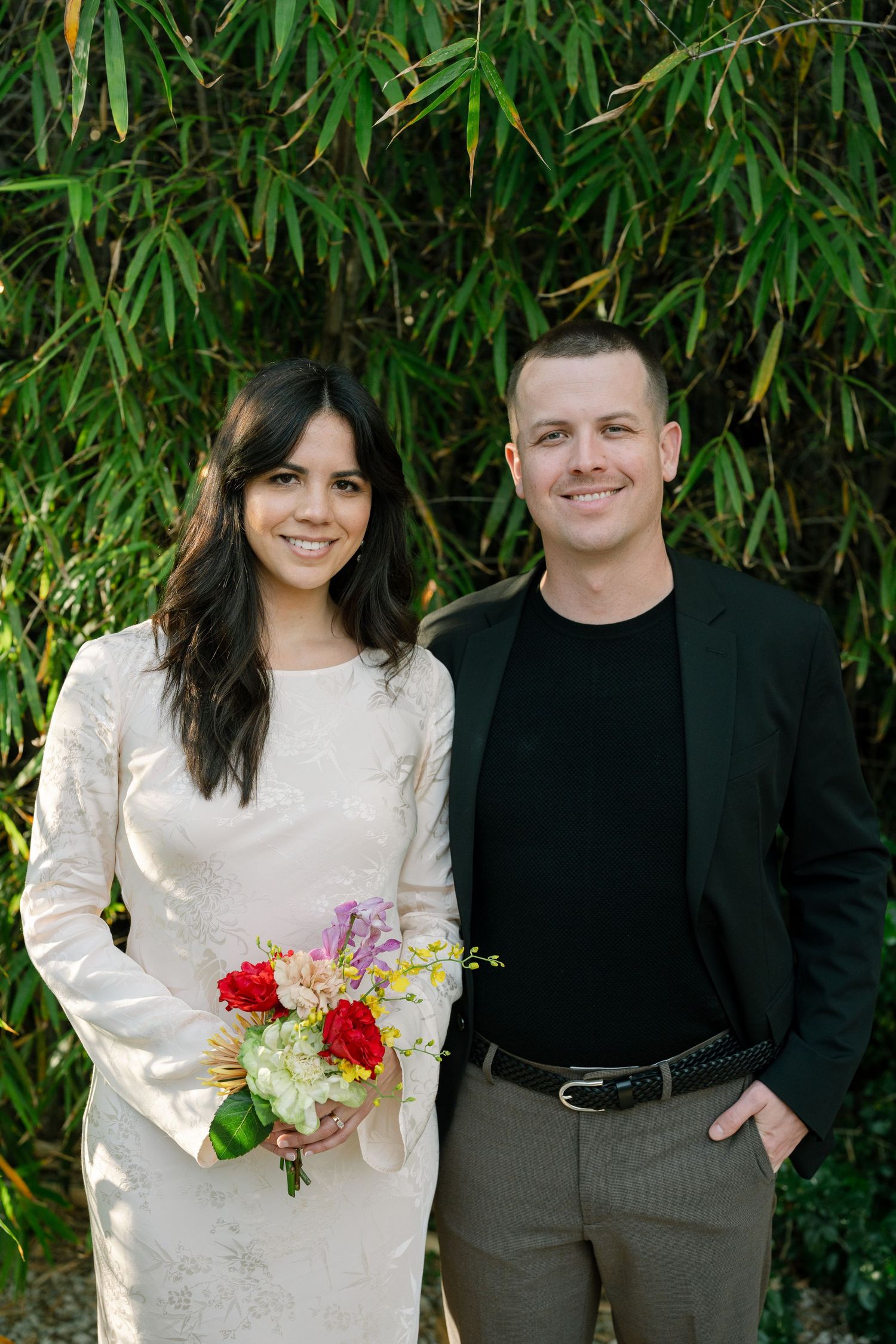Elegant couple in formal attire pose with bouquet against bamboo backdrop.