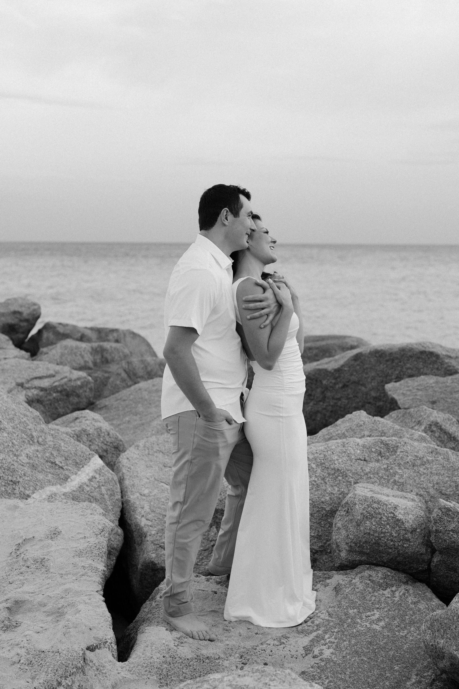 A couple in white clothing shares tender moments among large coastal rocks near the ocean.
