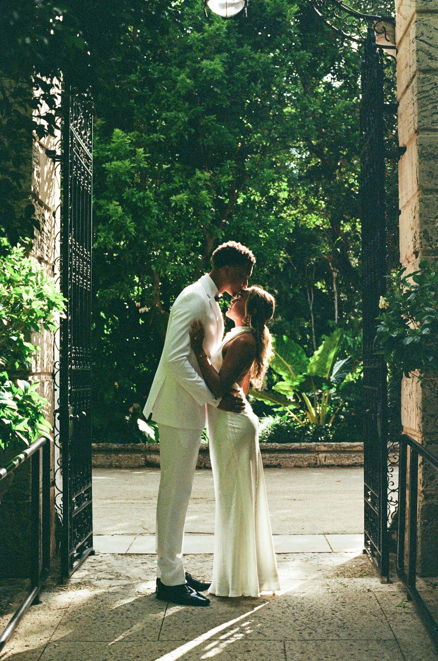 A romantic wedding couple shares an intimate moment in a sun-dappled forest setting with tall trees.