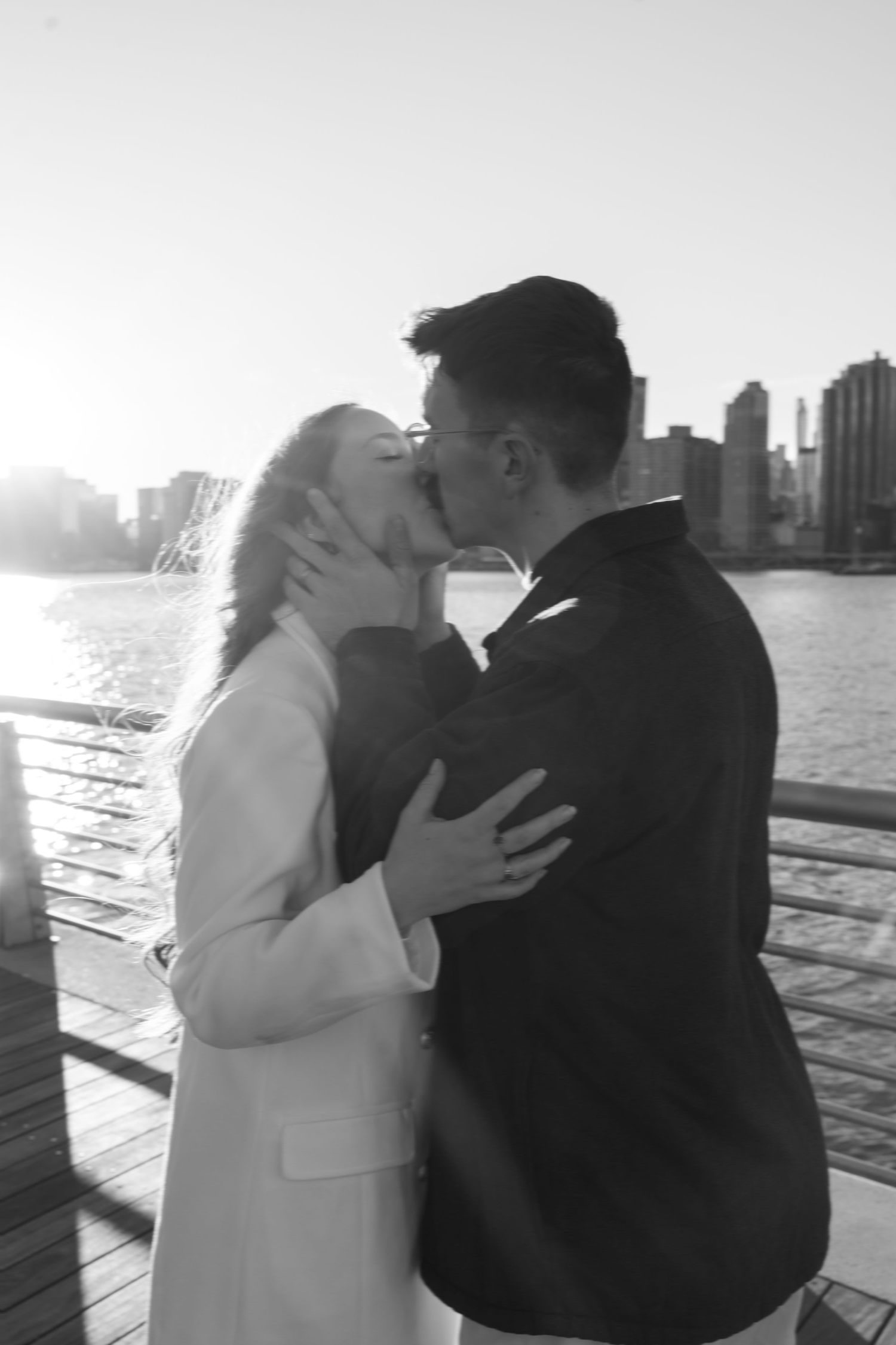 A couple shares an intimate moment at sunset on a waterfront boardwalk with a city skyline in the distance.