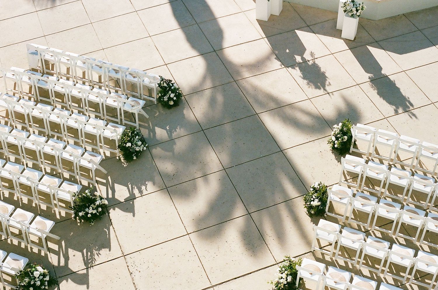Rows of white folding chairs arranged outdoors with green wreaths hanging on the end of each aisle for a wedding ceremony.