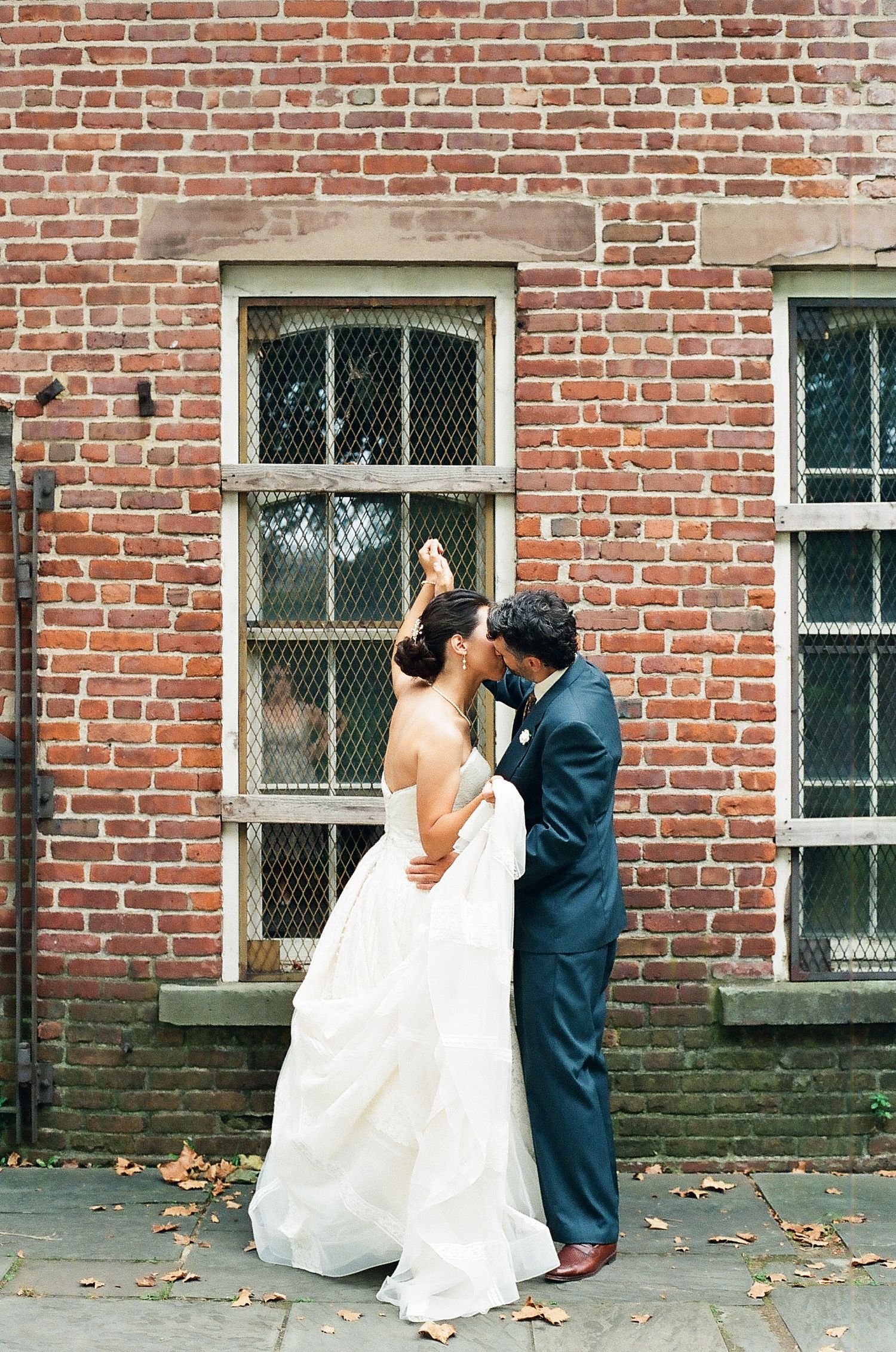 A romantic wedding couple embraces against a rustic brick wall with vintage windows.