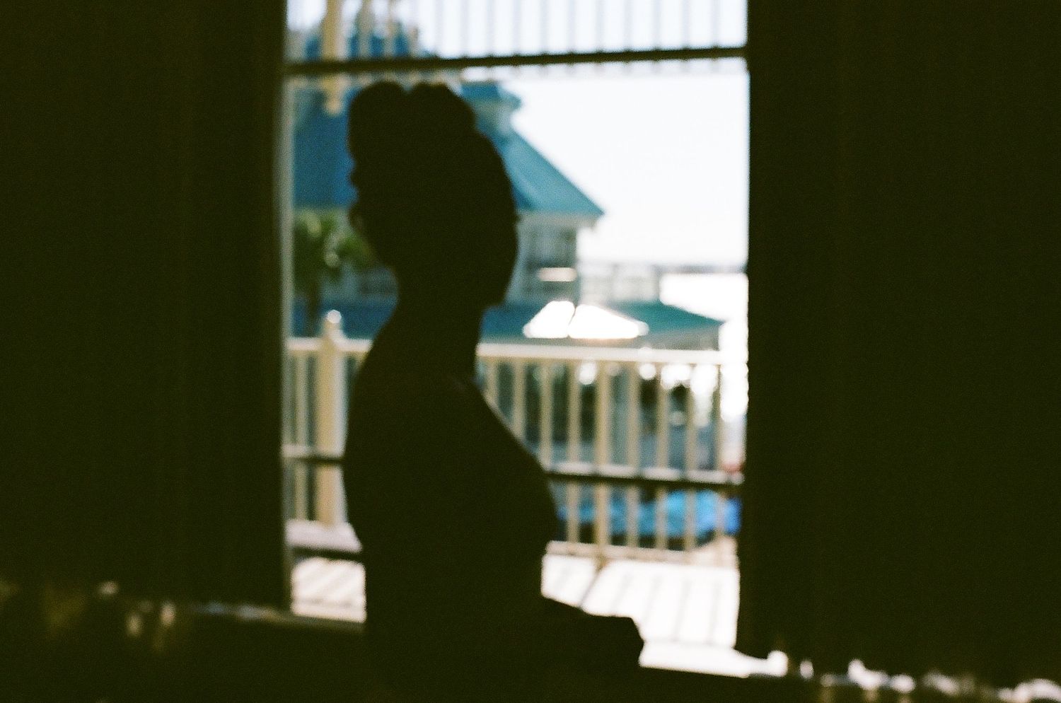 Silhouette of a person sitting on a bench against a bright window with white curtains and railings in view.