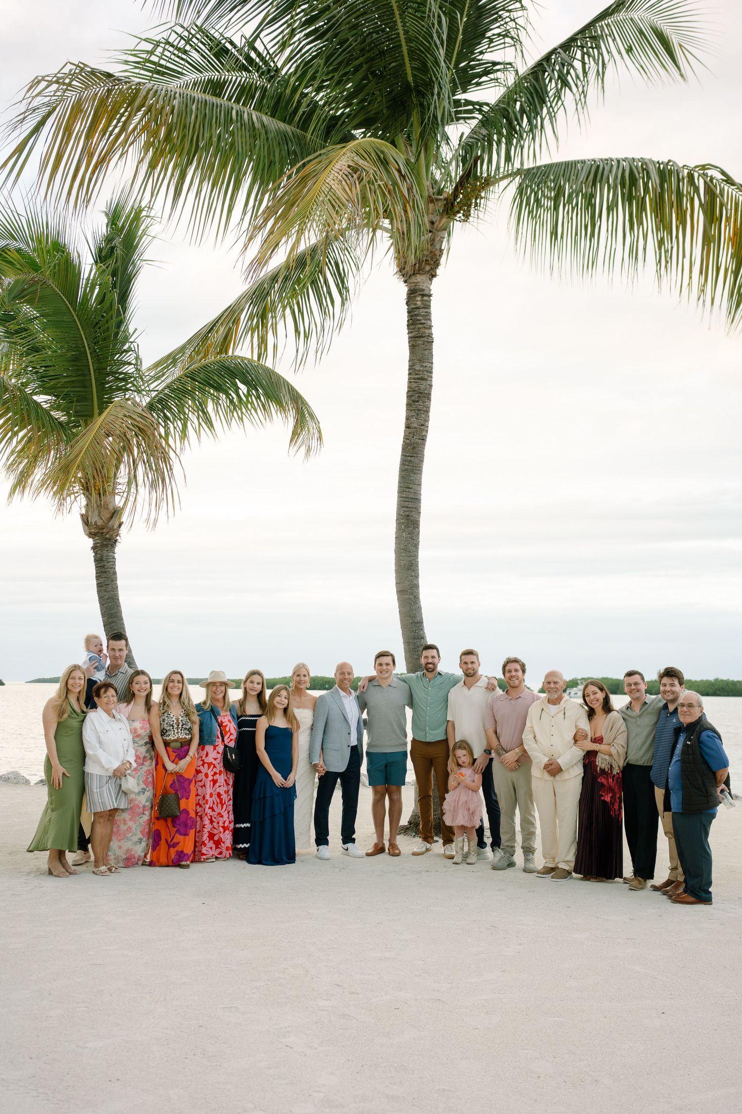 A large group of wedding guests pose on a tropical beach with palm trees and white sand.