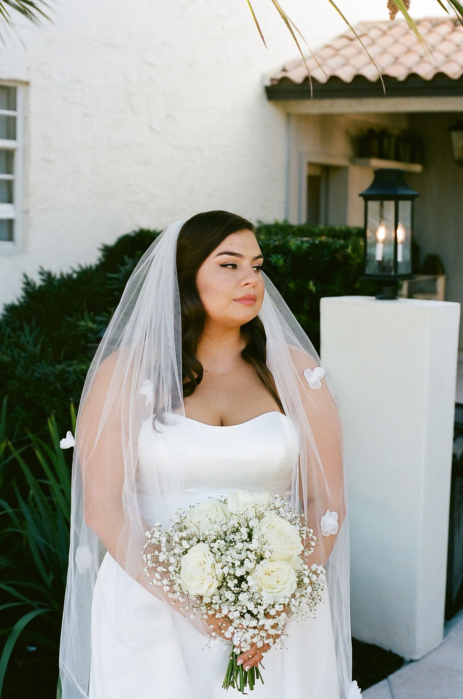 A bride in a white wedding dress holds a delicate bouquet of white roses and baby's breath against a light stucco wall.
