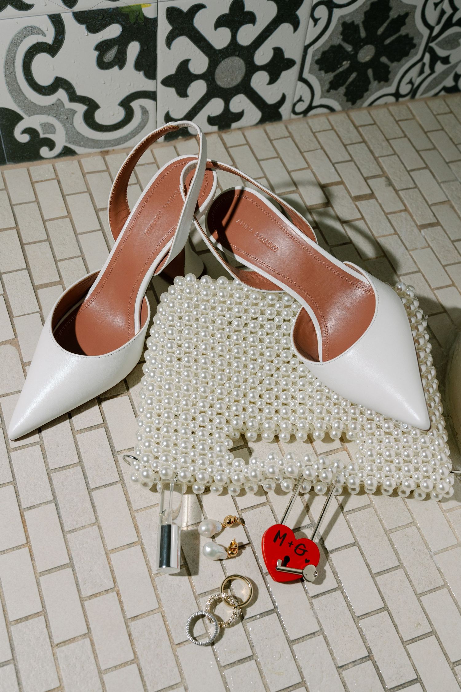 White slingback heels arranged with a pearl clutch purse, jewelry, and red heart charm on decorative tile flooring.