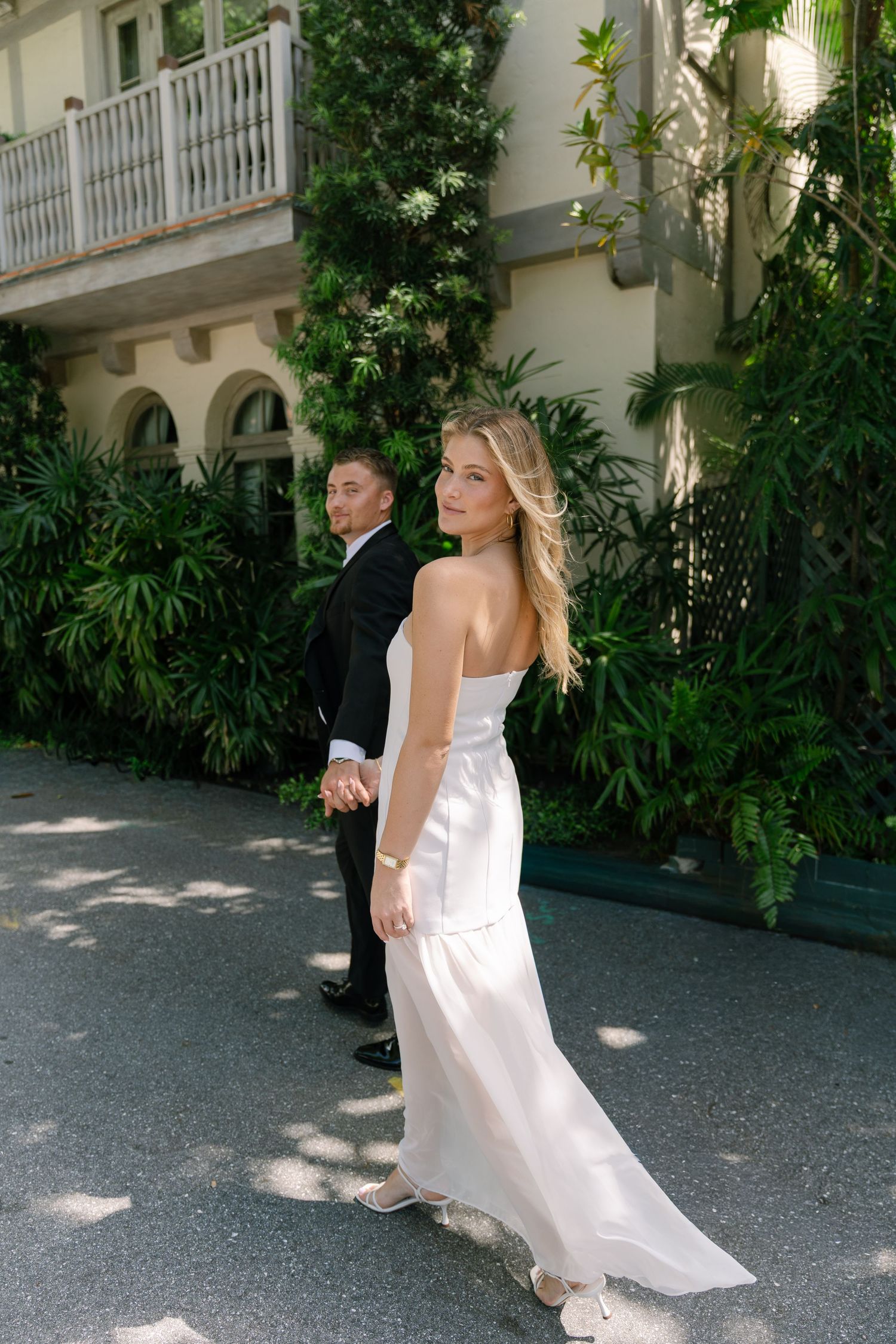 Bride in white gown holds groom's hand walking past lush greenery outside elegant building.