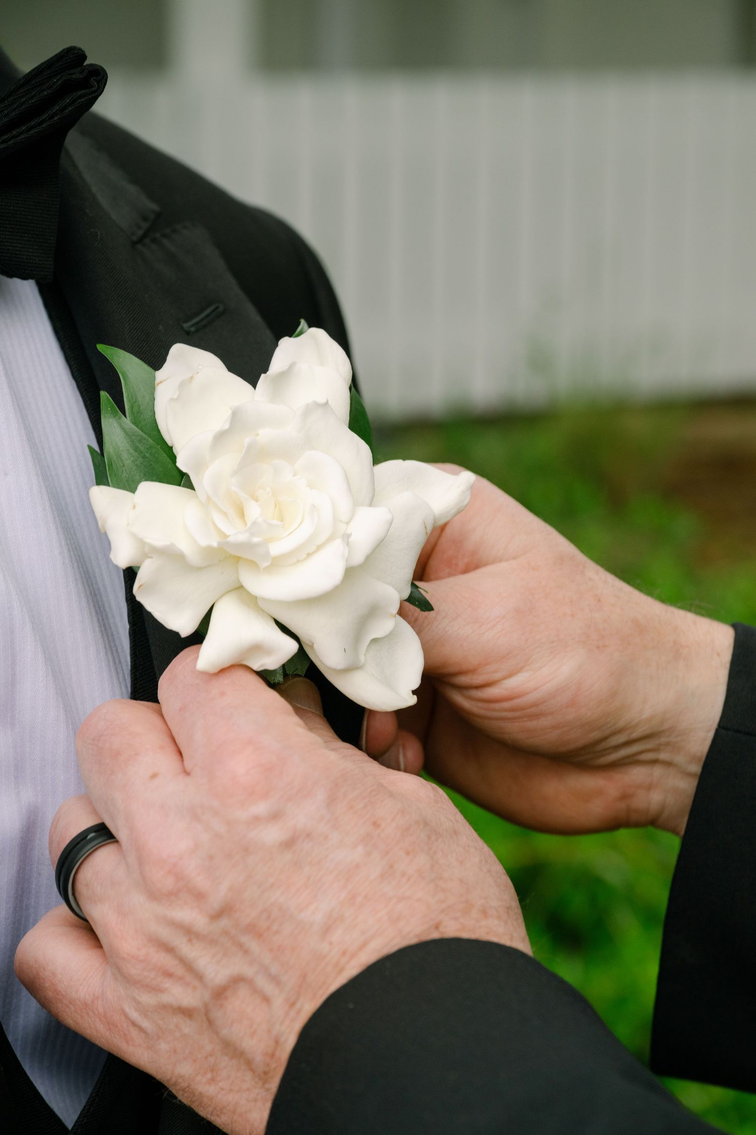 Hands pinning a white gardenia boutonniere onto a black tuxedo lapel at a wedding ceremony.