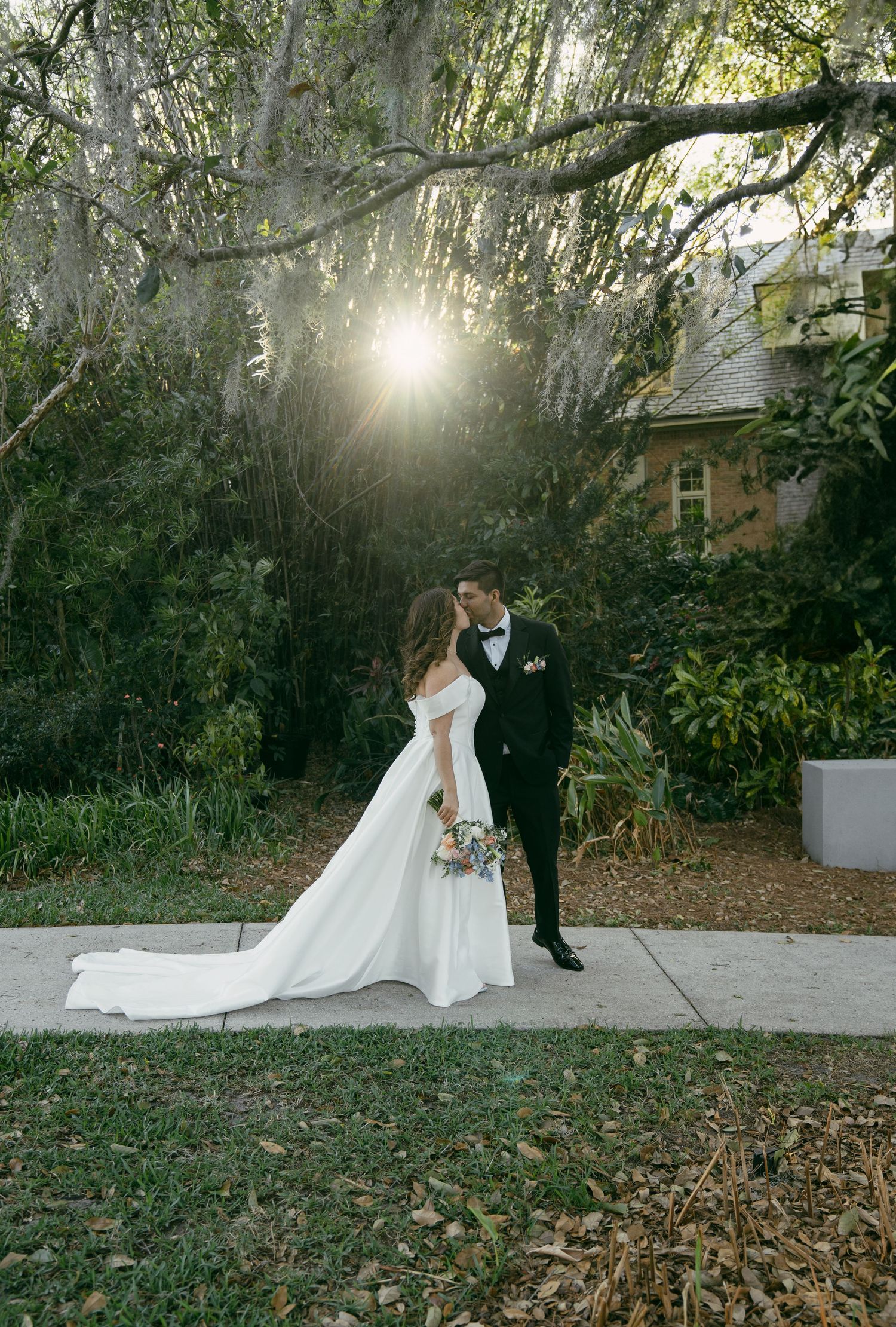 A wedding couple embraces in a scenic garden setting with sunlight streaming through Spanish moss-draped trees.