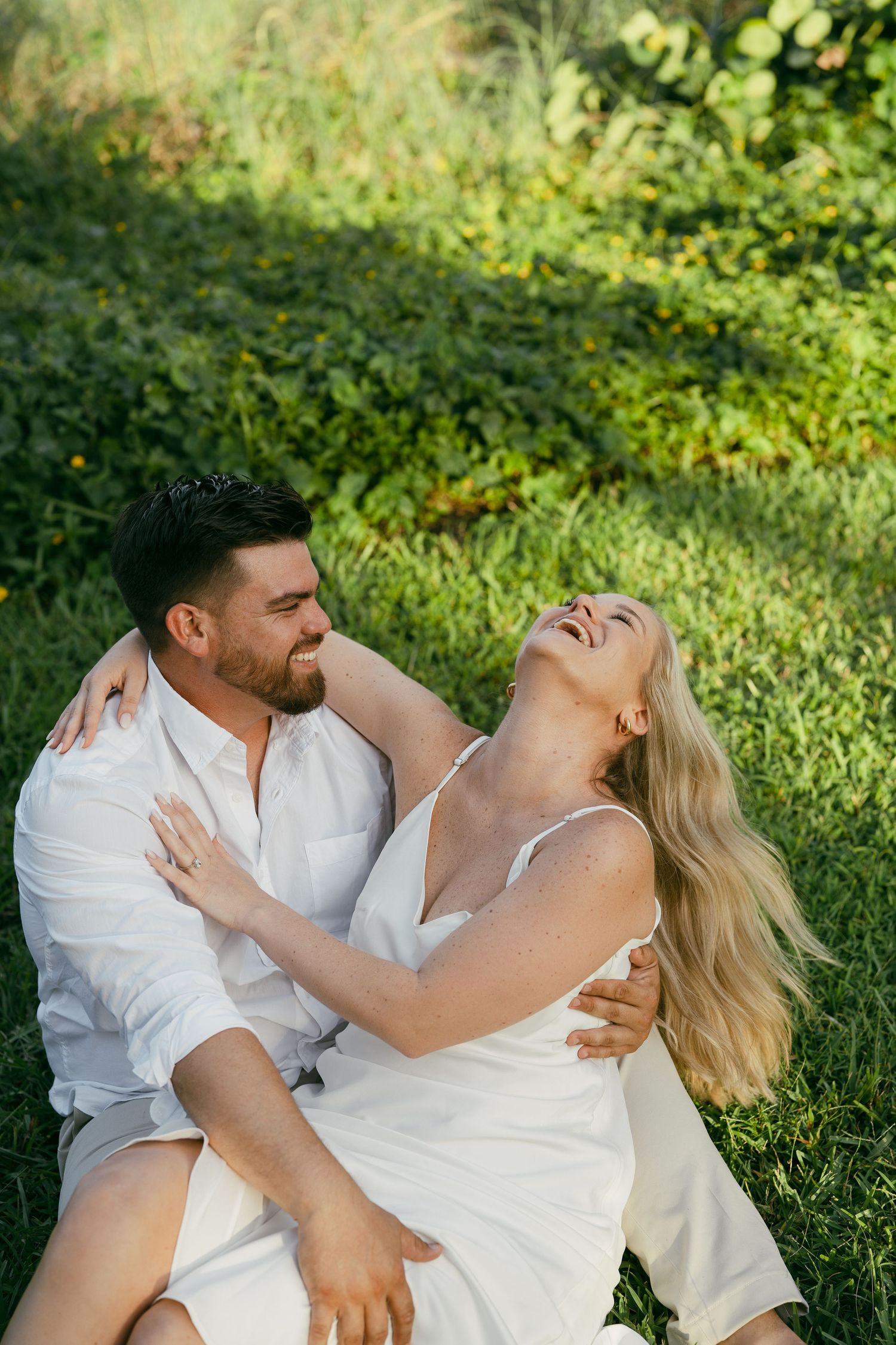 A couple in white outfits laughs together while sitting on green grass in a sunny garden setting.