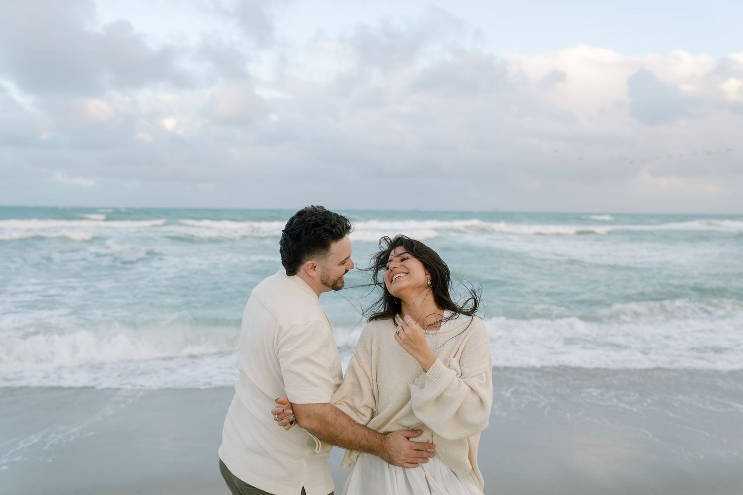 Two people share a romantic moment by the ocean shore during an overcast day.