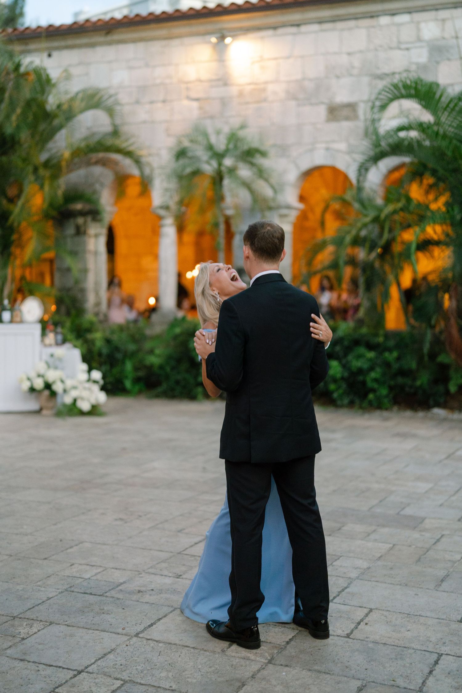 Bride in blue gown and groom in black tuxedo sharing romantic first dance outside illuminated stone venue at dusk.