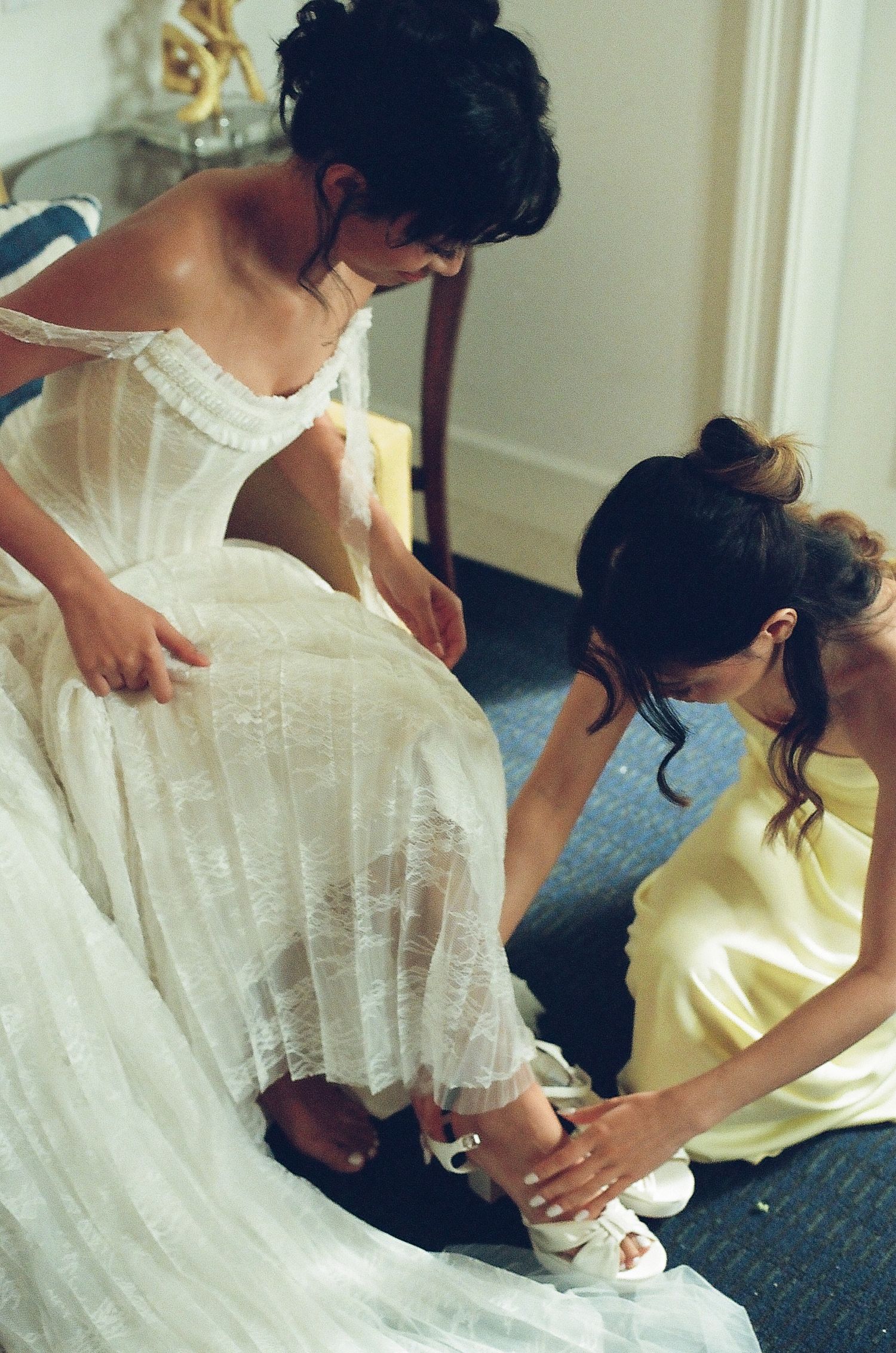 Bridesmaid in yellow satin dress helps bride put on white heeled sandals while another assists with wedding gown.