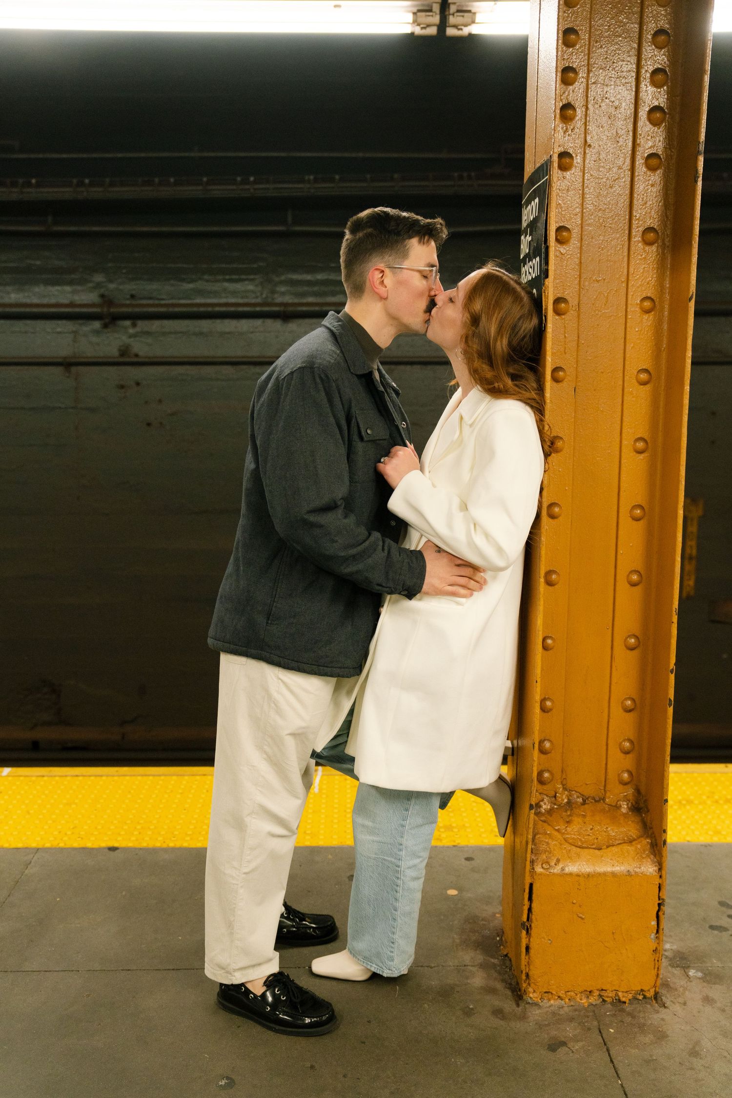 A couple shares an intimate moment by a yellow industrial pillar in an urban setting.