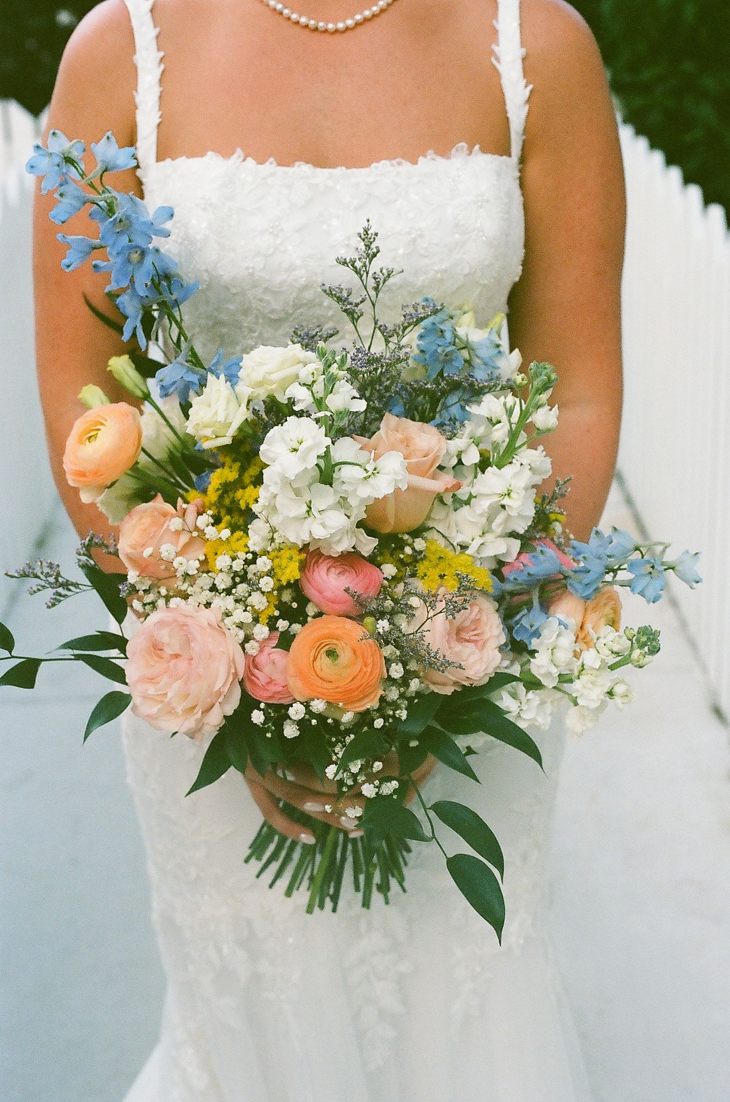 Romantic bridal bouquet featuring peach ranunculus, blue delphinium, and white baby's breath against white wedding dress.