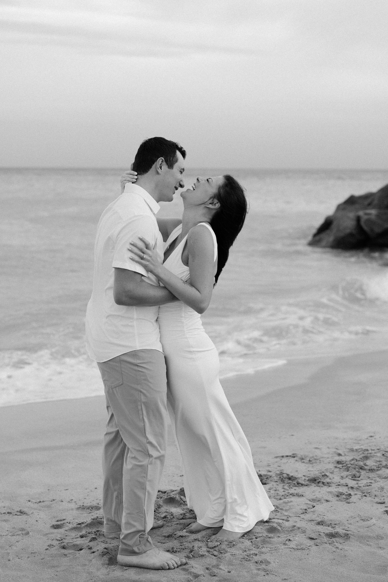 A romantic black and white beach portrait of a couple embracing on the sand with ocean waves in the background.