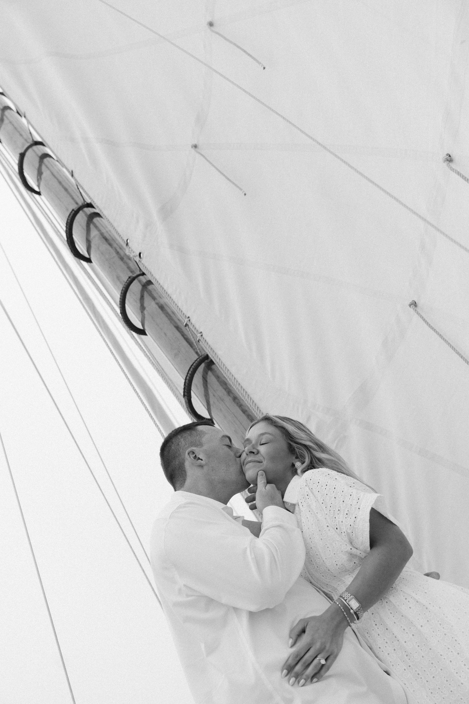 Black and white photo of a couple sharing a romantic kiss on a sailboat with mast and rigging above.