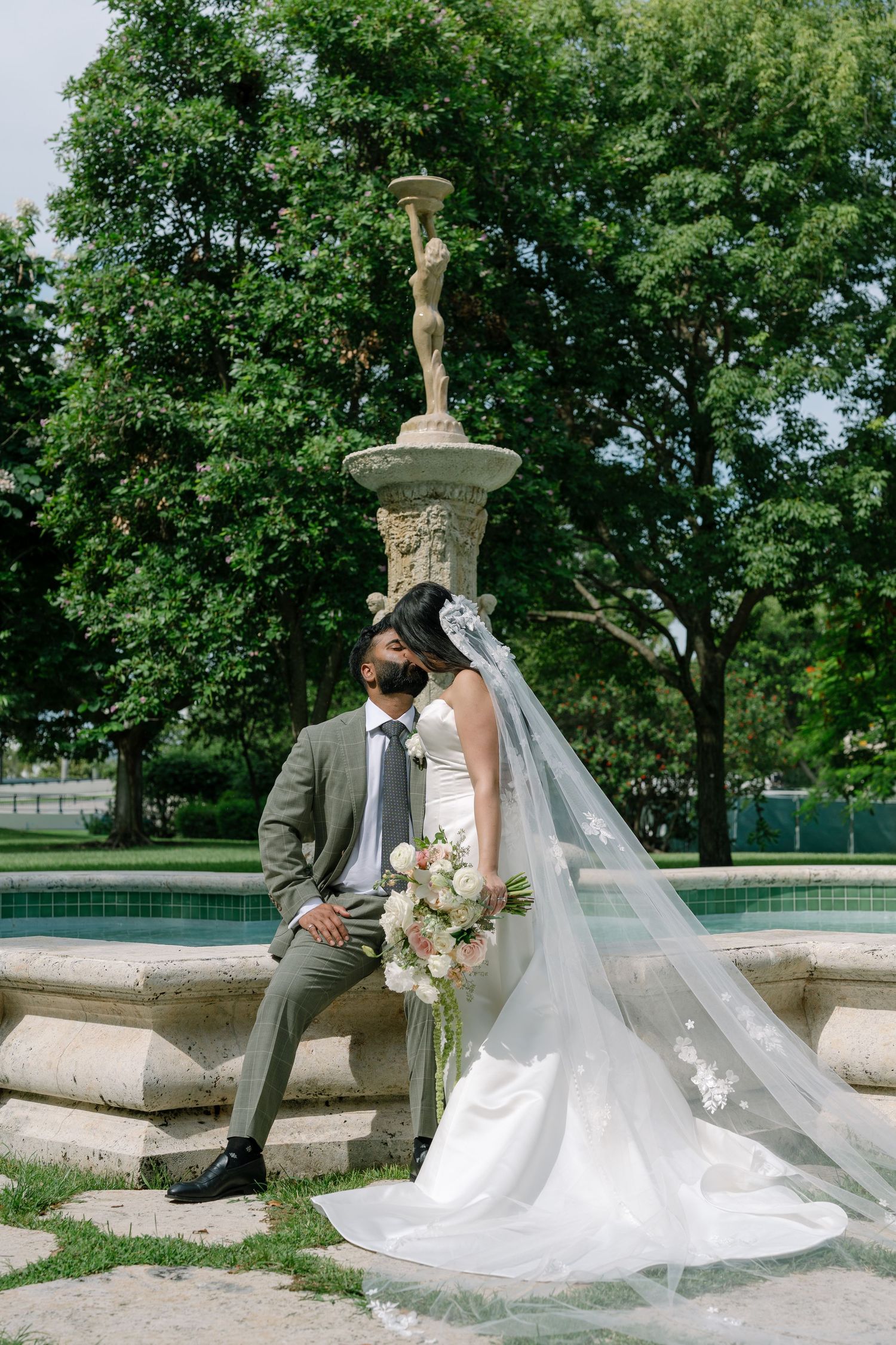 Bride and groom share a romantic moment by a stone fountain in a lush garden, her veil flowing gracefully.