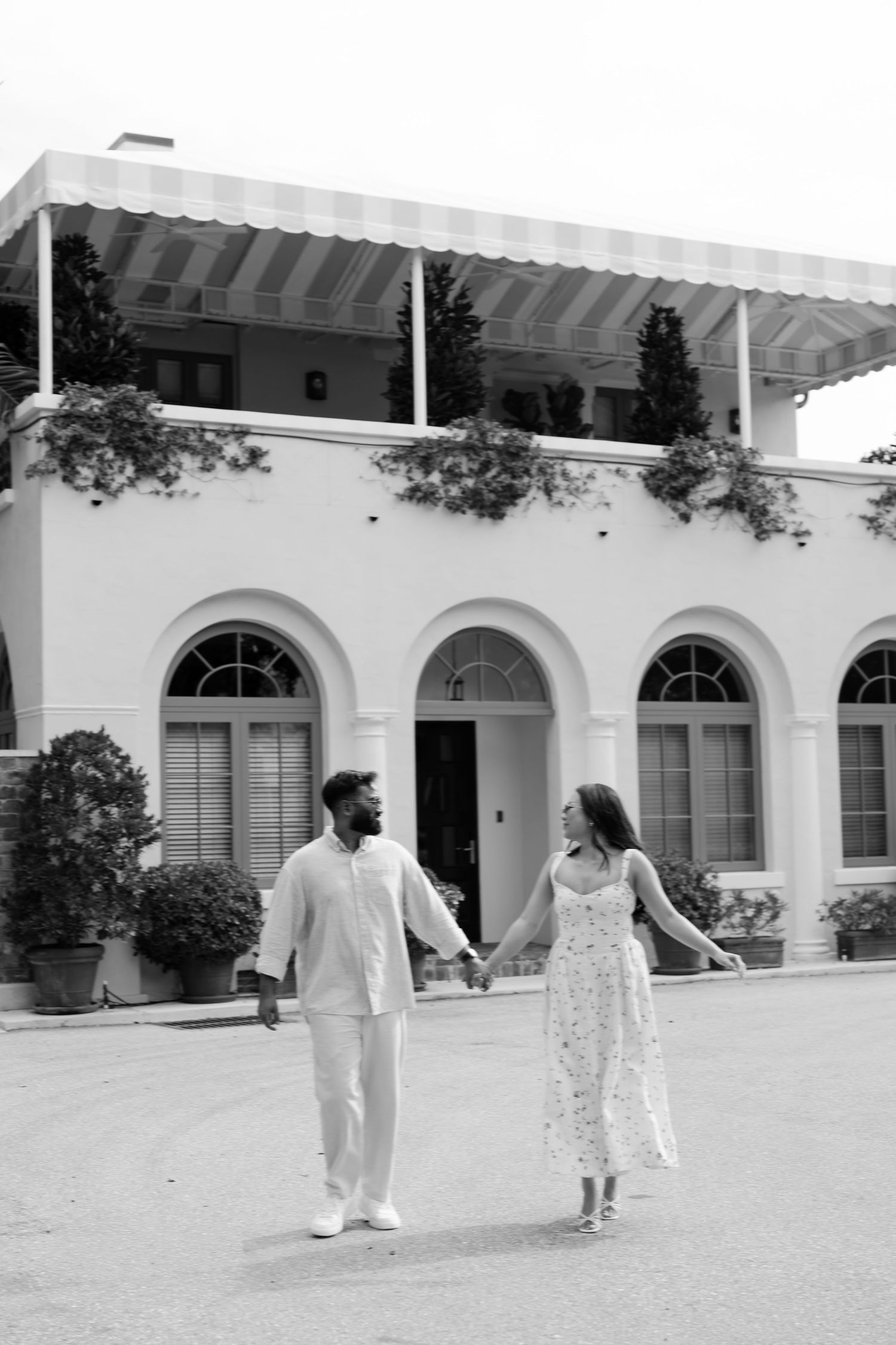 Black and white photo of a couple holding hands in front of an elegant white Mediterranean-style villa.