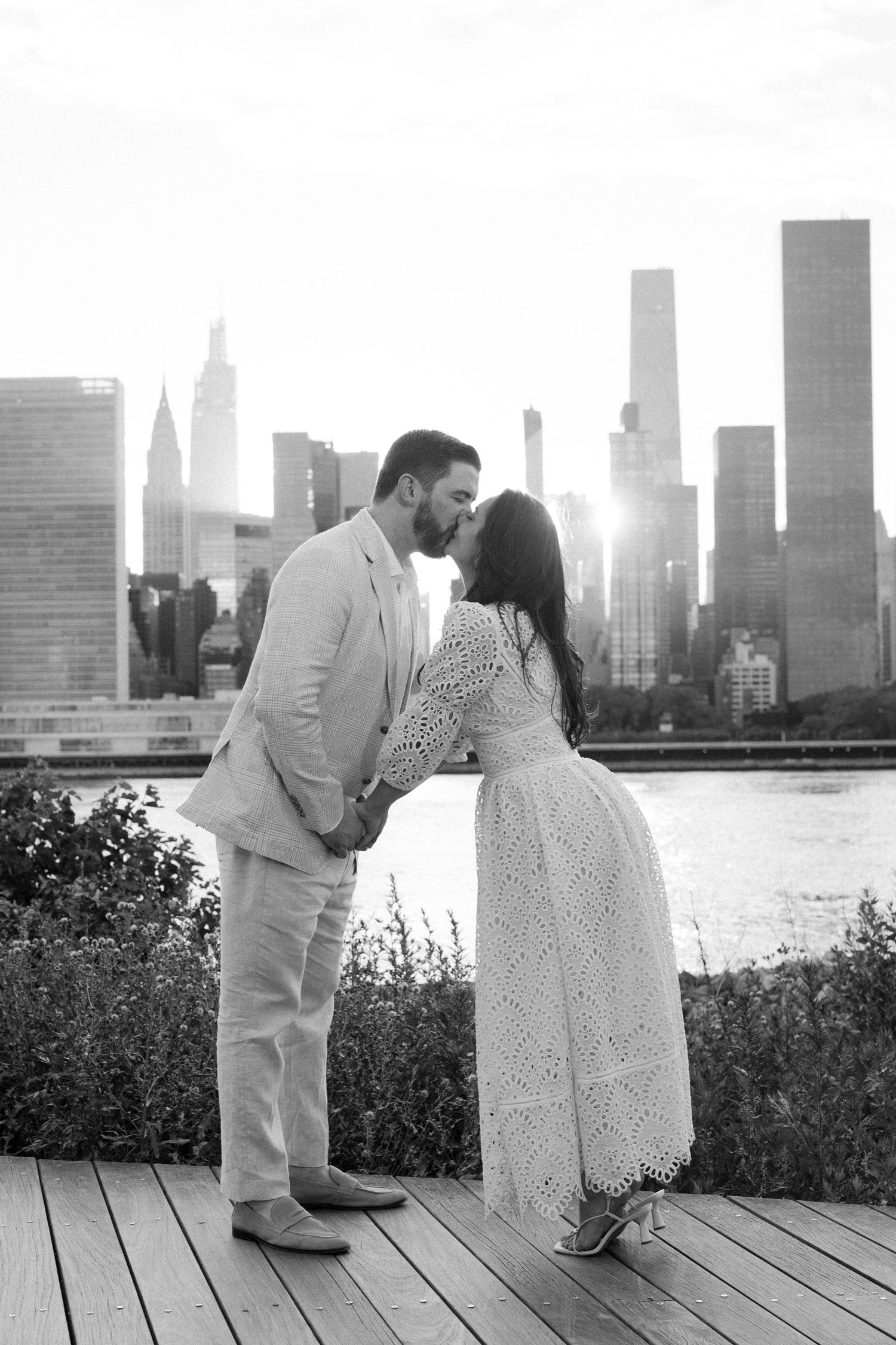 Black and white photo of a couple kissing on a wooden deck with NYC skyline and river in the background.