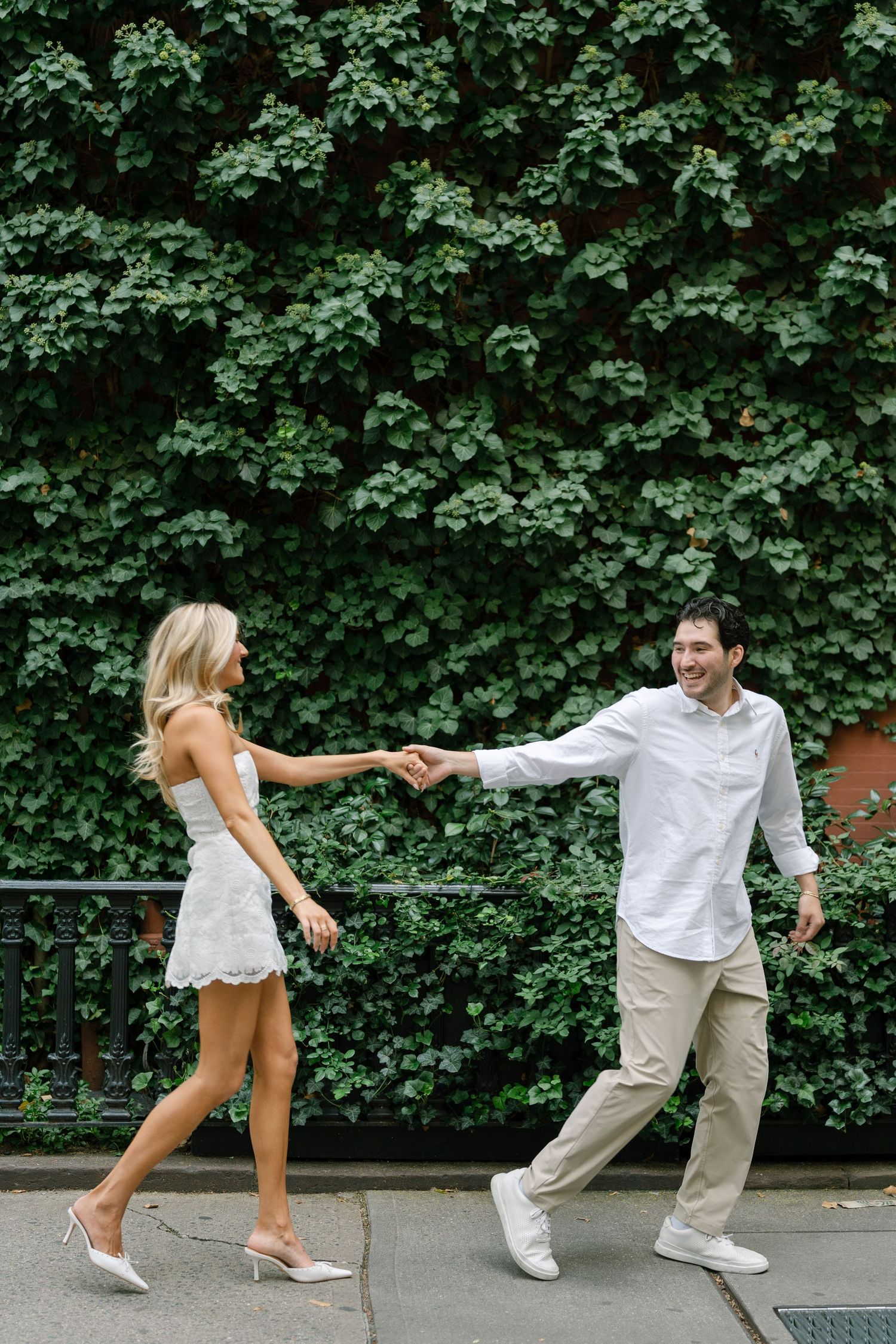 Smiling couple holding hands and walking along a sidewalk covered by a lush green ivy-covered wall.