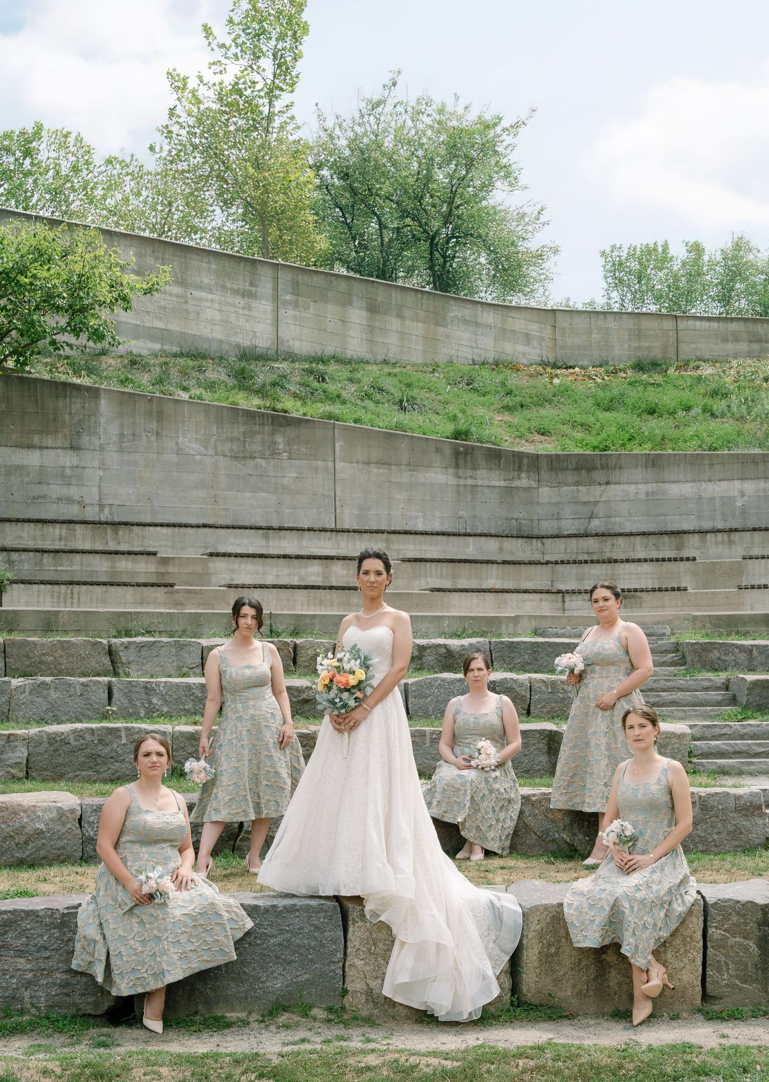 A wedding party in gray bridesmaid dresses and white bridal gown pose casually on stone amphitheater steps outdoors.