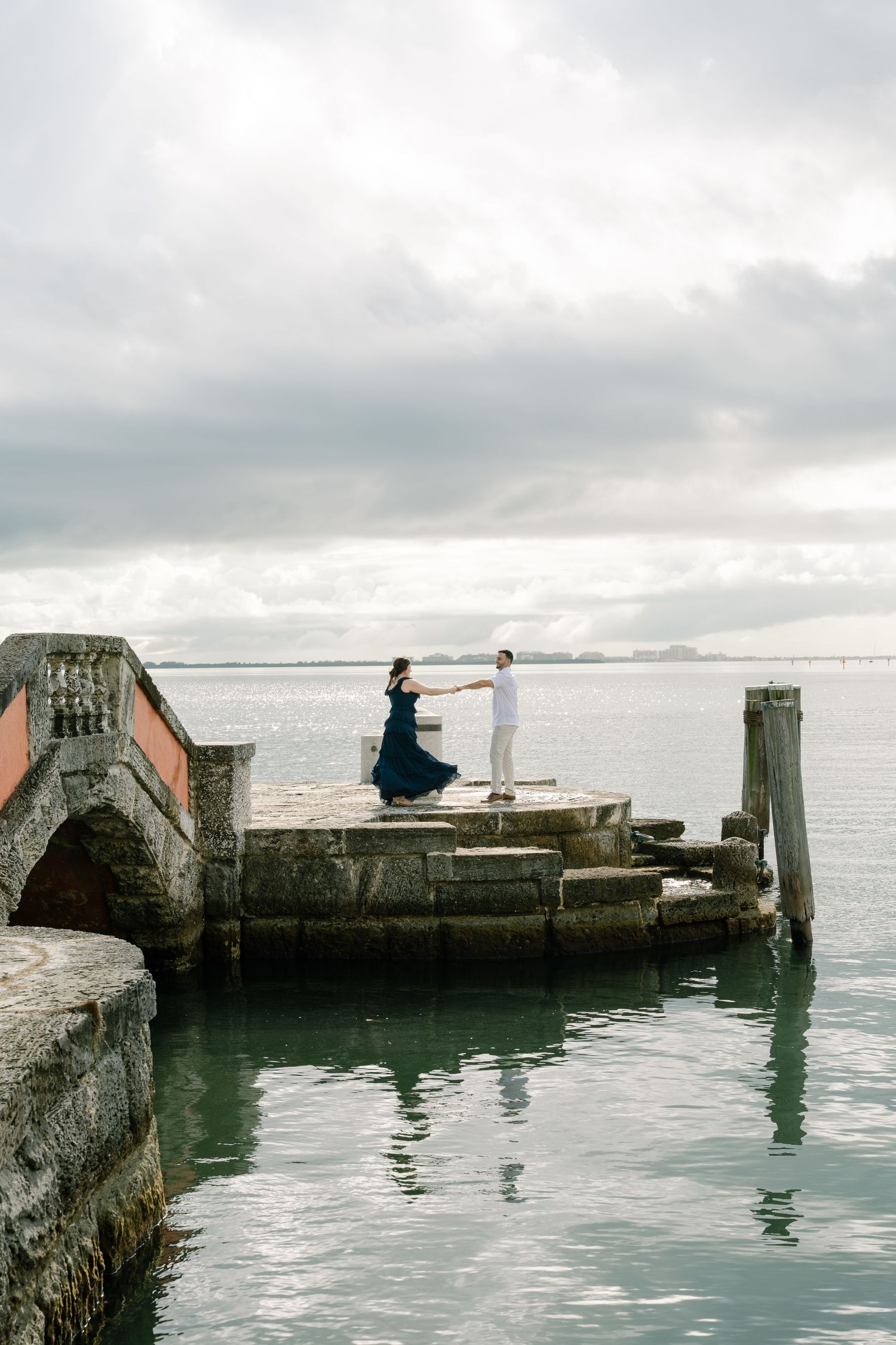 A couple shares a romantic moment on a seaside pier with cloudy skies and ocean waves in the background.