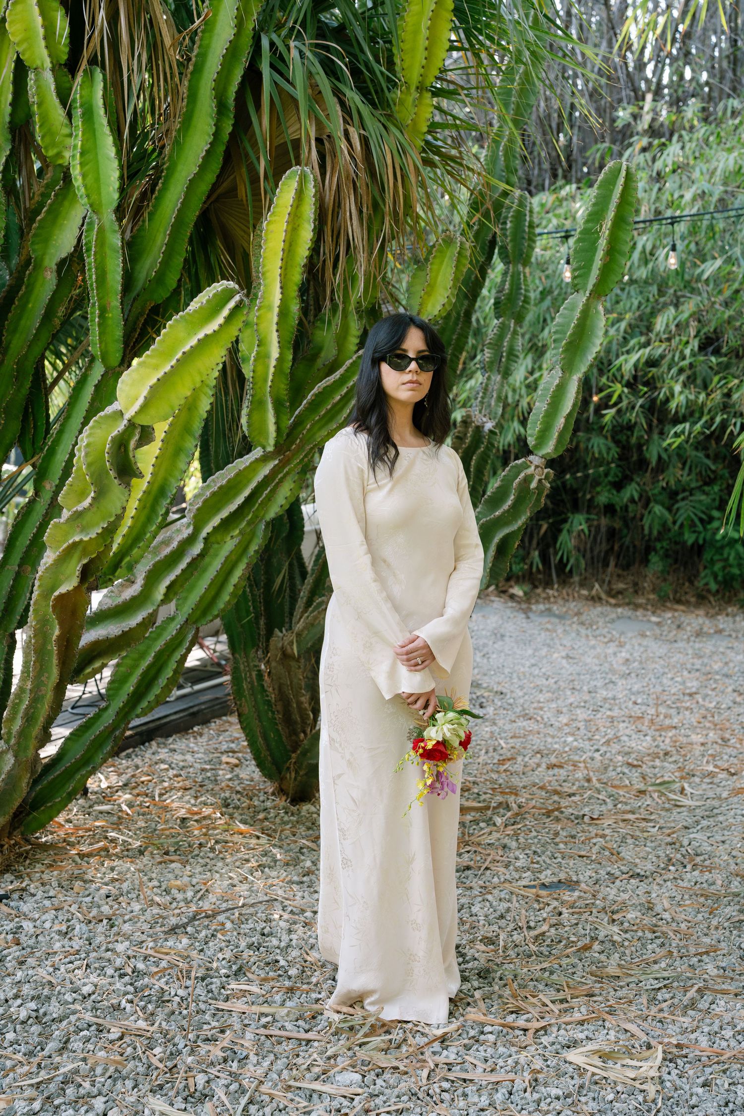 A figure in a flowing cream dress poses against a backdrop of tall cacti and tropical plants in an outdoor garden setting.