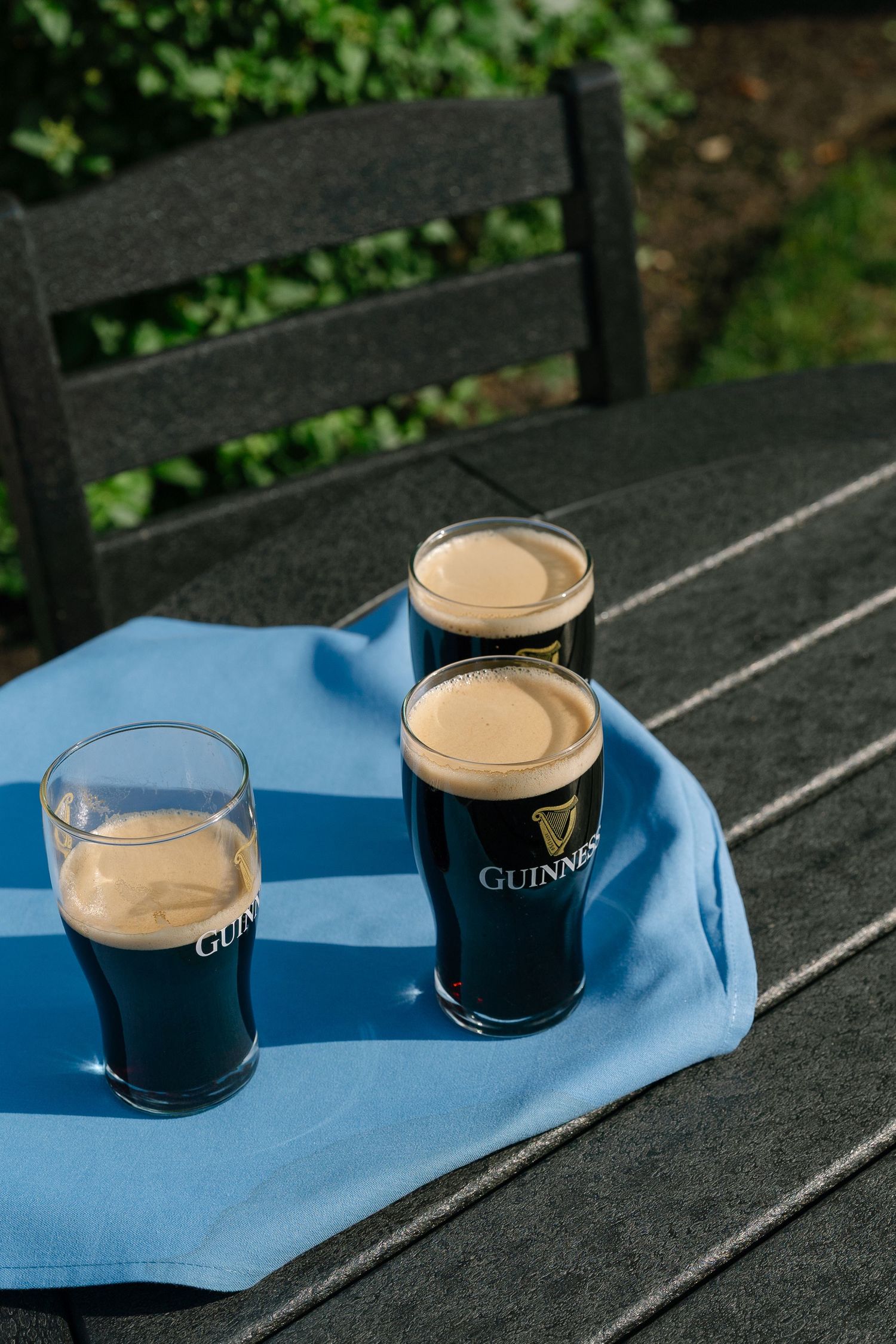 Three pints of Guinness stout on a blue cloth on a dark outdoor table, with creamy heads and lush greenery behind.