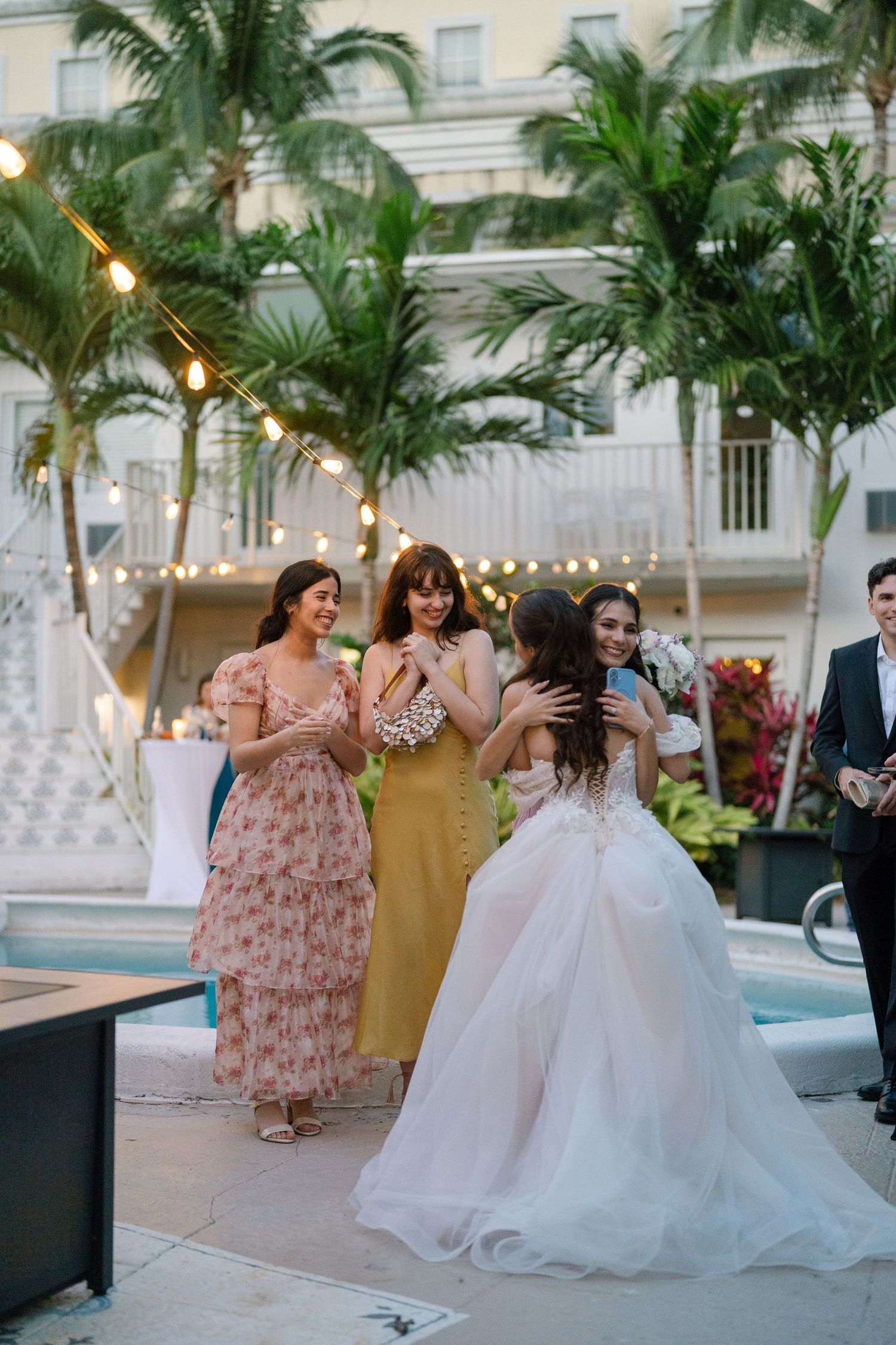 Bride in white ball gown greeted by bridesmaids in colorful dresses at outdoor wedding reception.