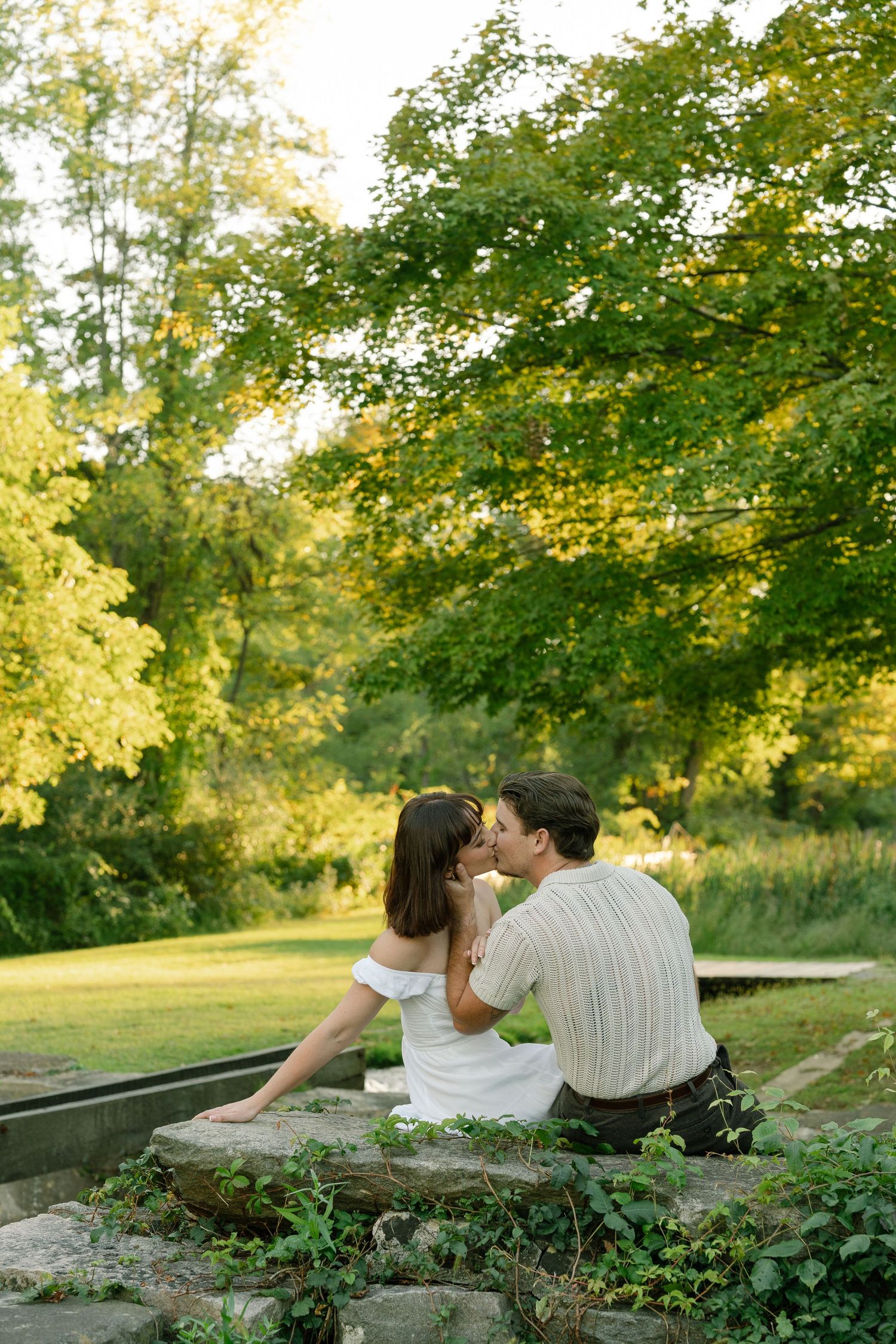 Romantic couple sharing a kiss while sitting on stone steps surrounded by vibrant green trees in a park.
