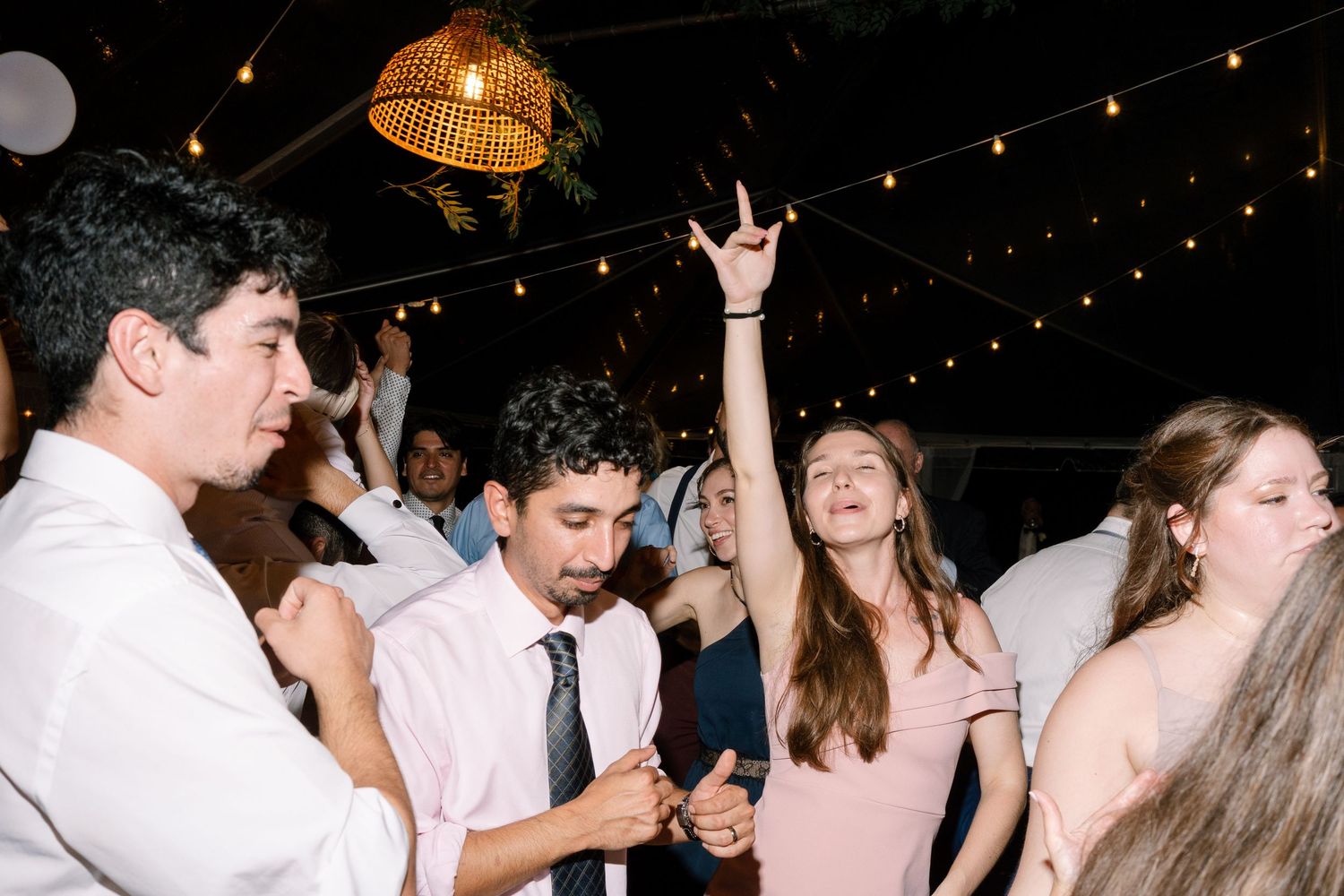 Energetic wedding guests dancing under string lights and disco ball at lively indoor reception.
