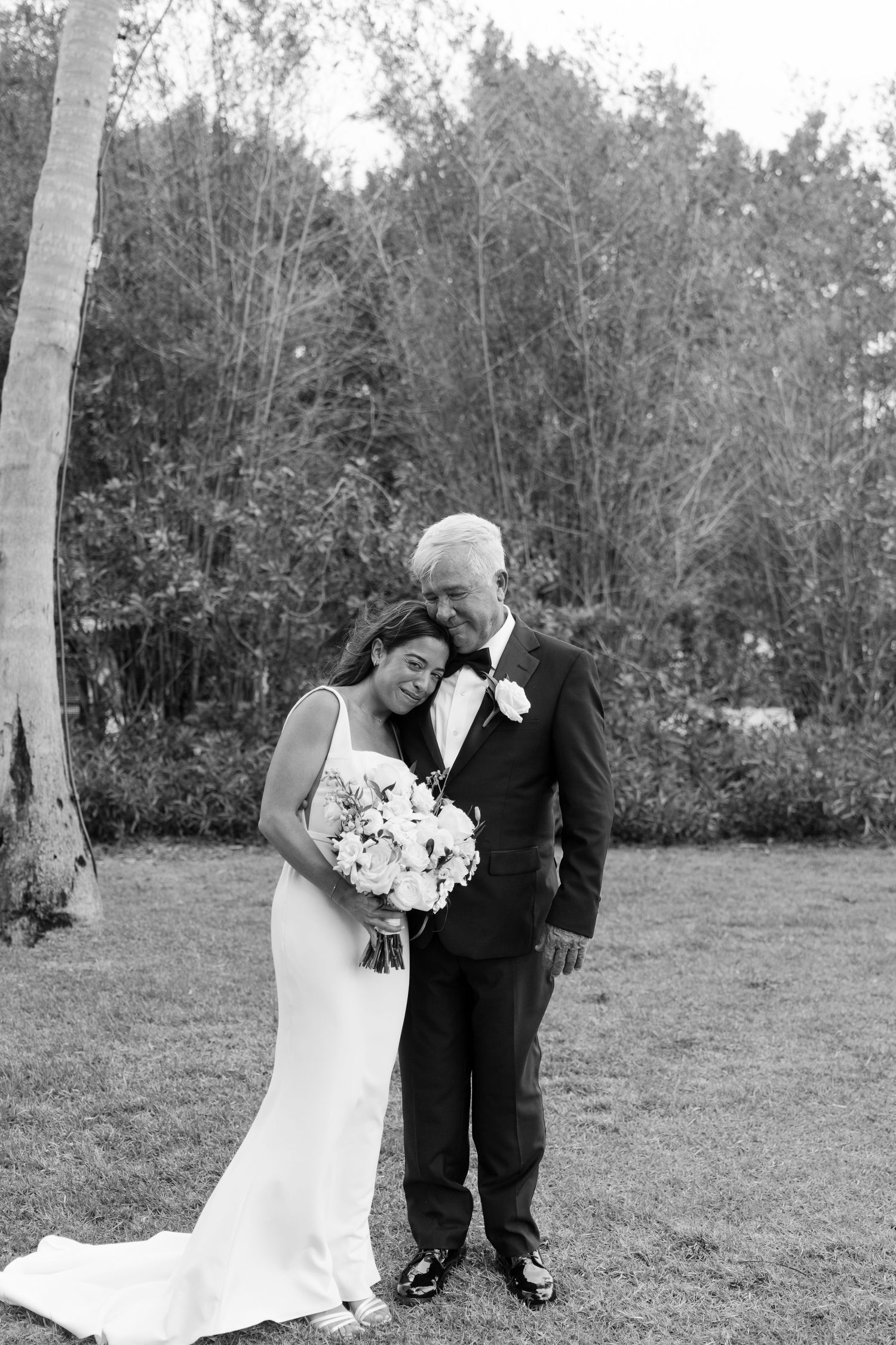 Black and white photo of a bride and groom embracing outdoors on grass, bride holding white bouquet, trees in background.